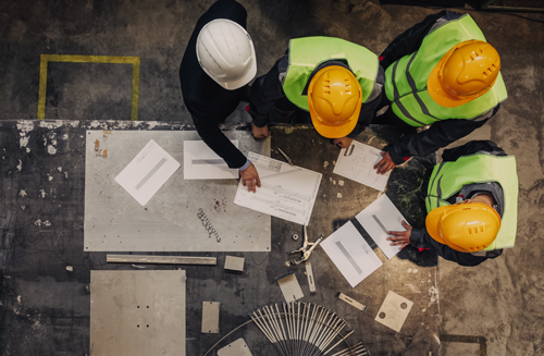 Four construction workers wearing yellow hard hats and high-visibility vests reviewing architectural plans and blueprints on a concrete floor, surrounded by construction materials and tools.