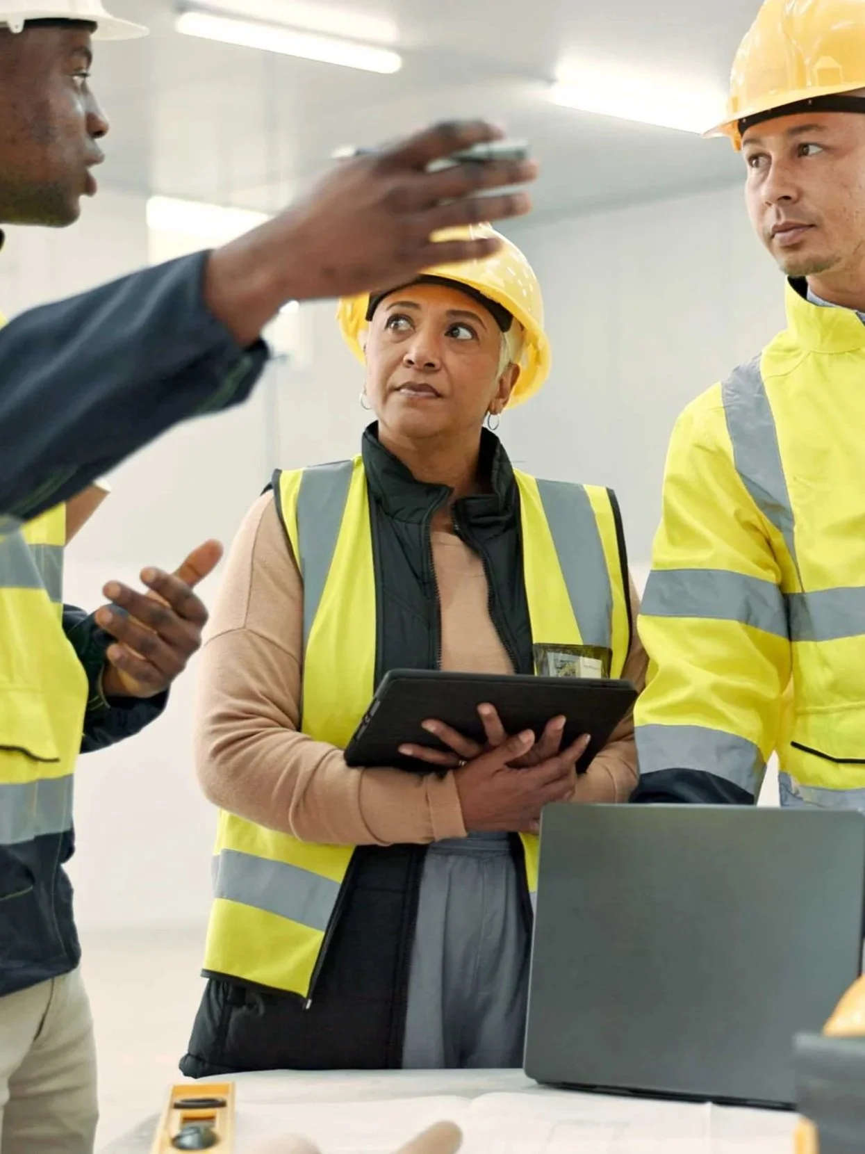 Group of construction workers wearing yellow safety vests and helmets, engaged in a discussion indoors with a woman holding a tablet.