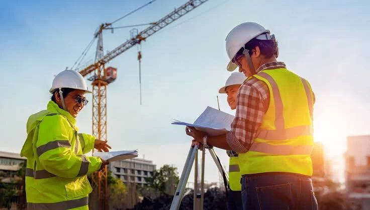 Group of construction workers wearing safety helmets and reflective vests discussing plans at a construction site with a crane in the background during sunset.