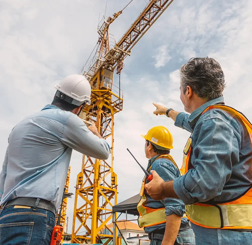 Three construction workers wearing safety helmets and vests are discussing a tall yellow crane at a construction site during daytime.