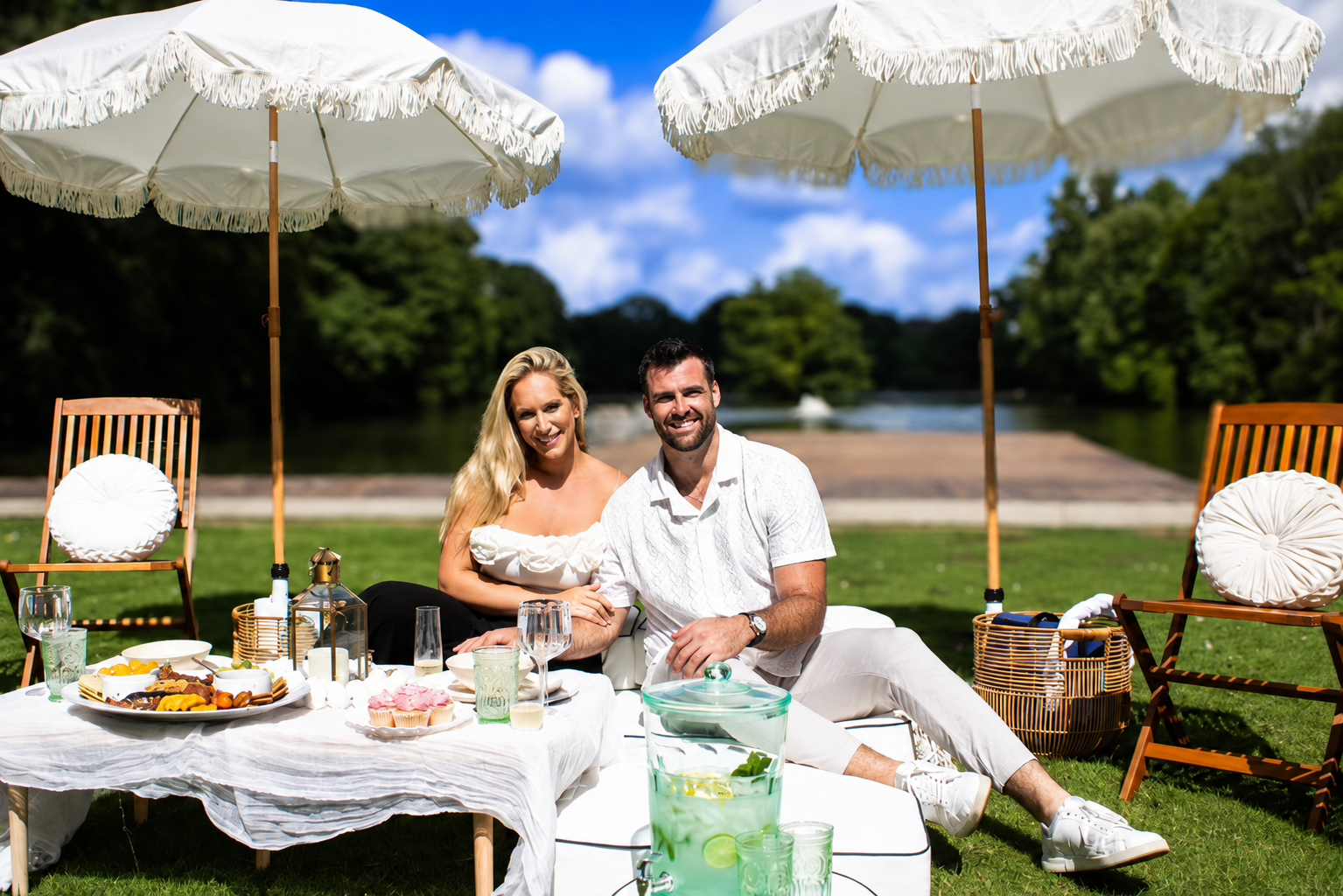 A couple enjoying a picnic outdoors under large white umbrellas, sitting at a table with assorted snacks and drinks, with a river and green trees in the background on a sunny day.