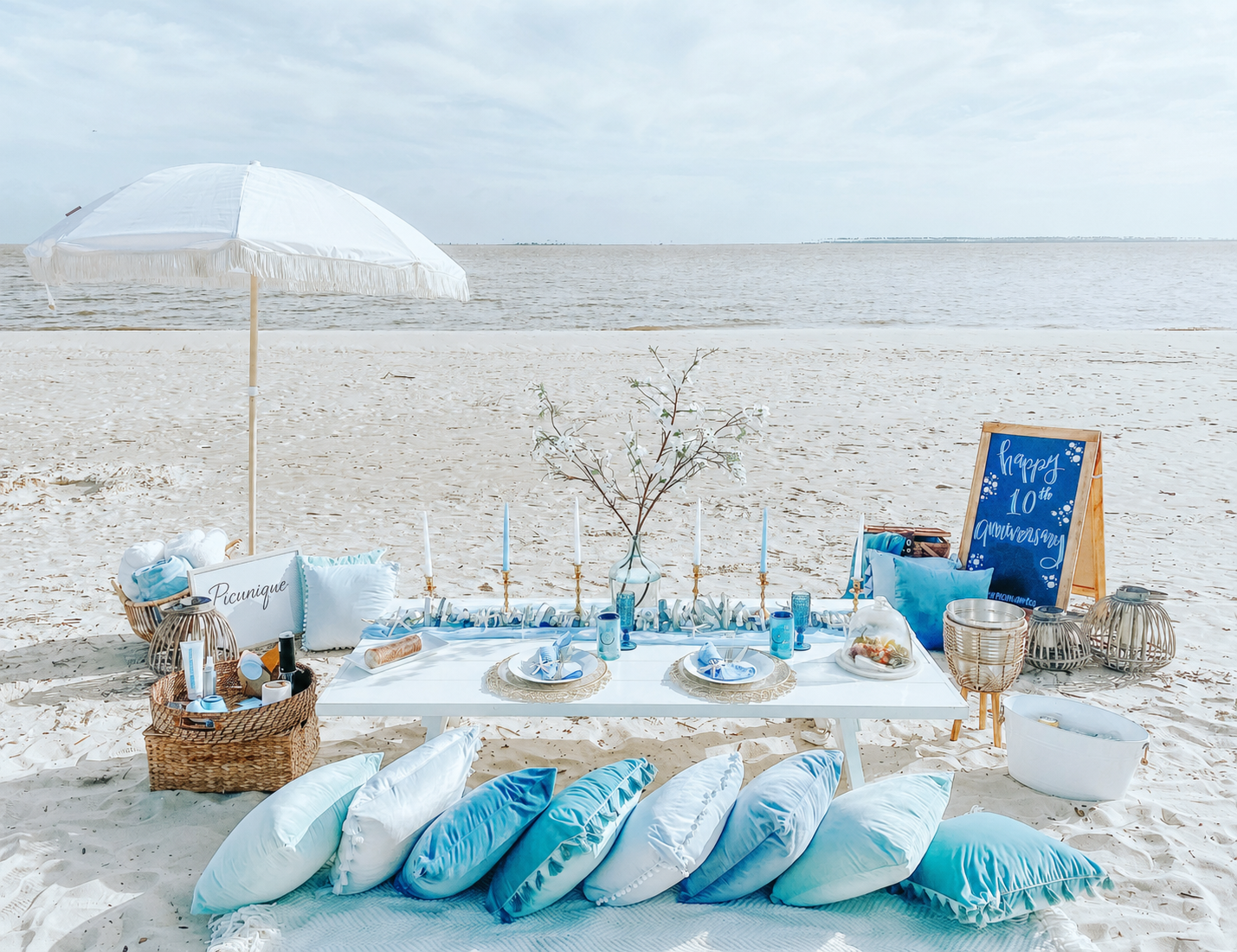 Beach setup for a celebration with a picnic table, pillows, candles, a sign, and decorations on the sand near the water.