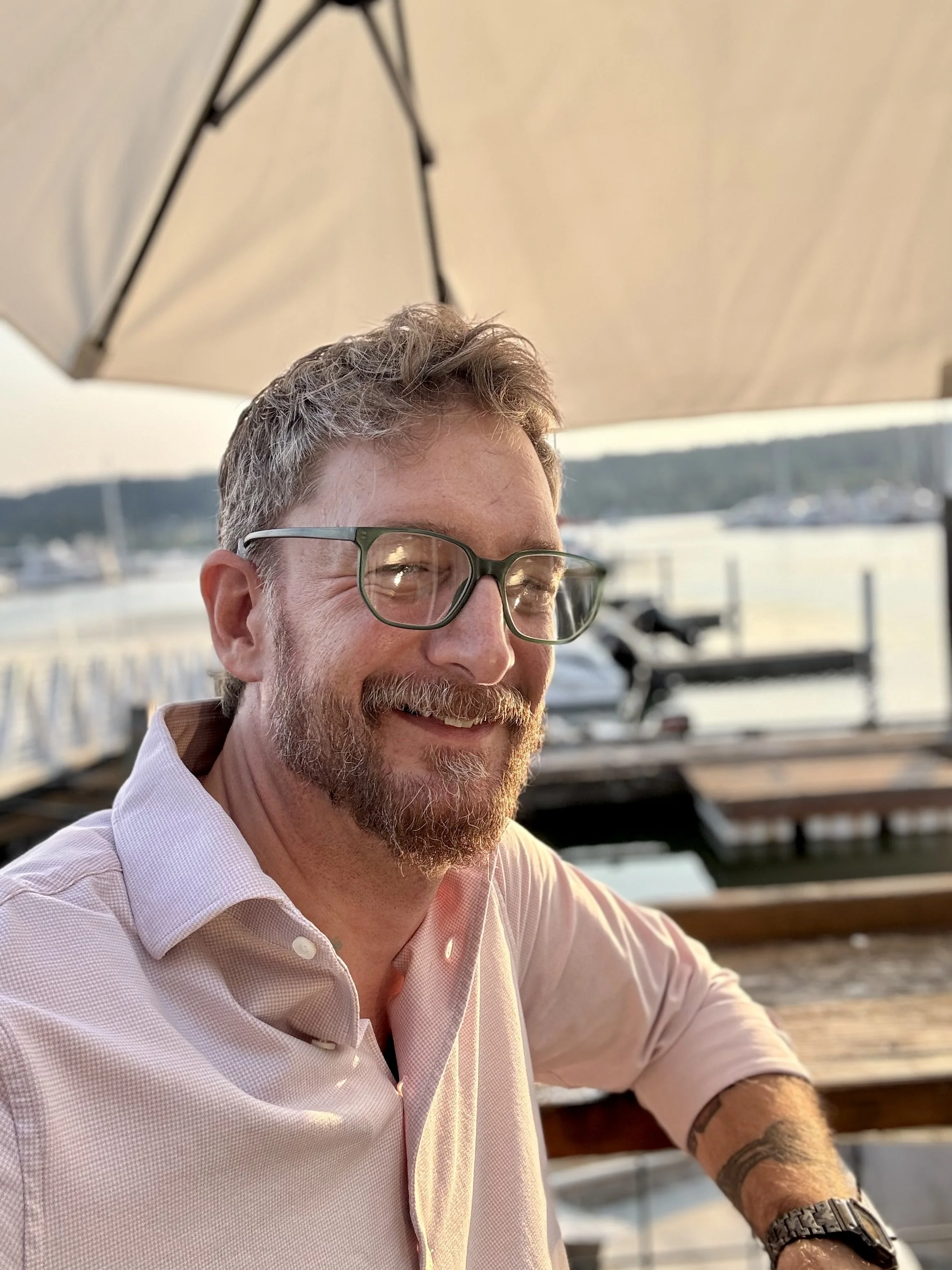 J. Michael "Mikey" Decker, leadership and career coach, smiling outdoors near a marina at sunset with boats in the background.