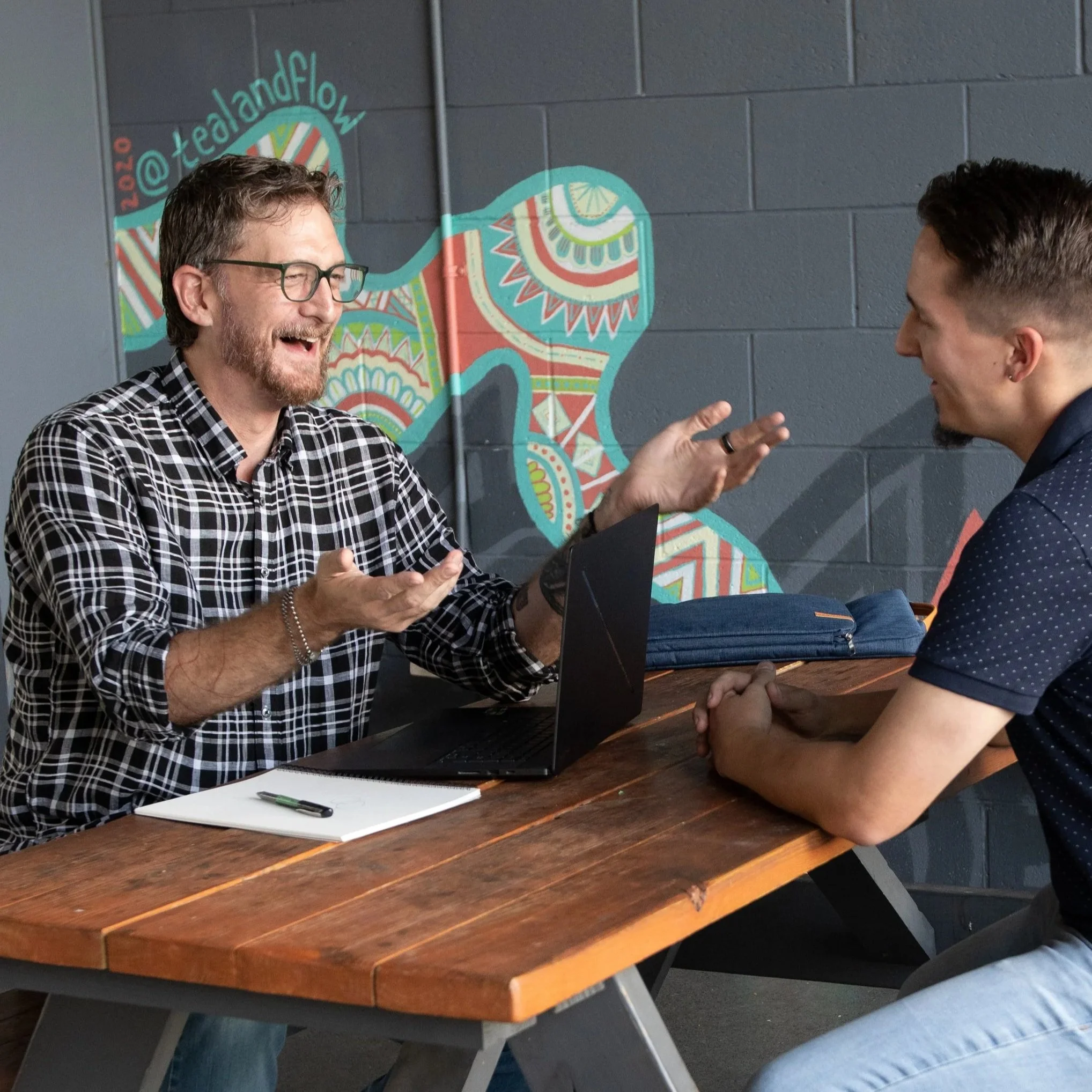 J. Michael Decker (Mikey) gesturing as he talks with a client across a table, a mural on the wall behind them.