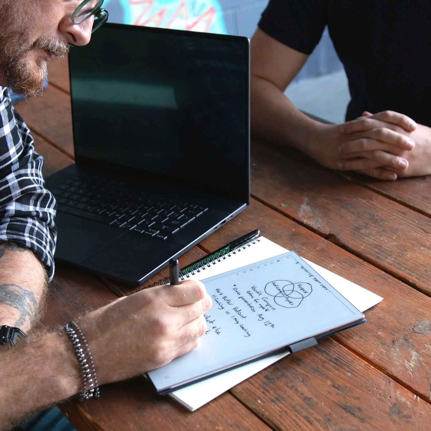 Mikey's hands sketching notes in a notebook beside a laptop on a wooden table.