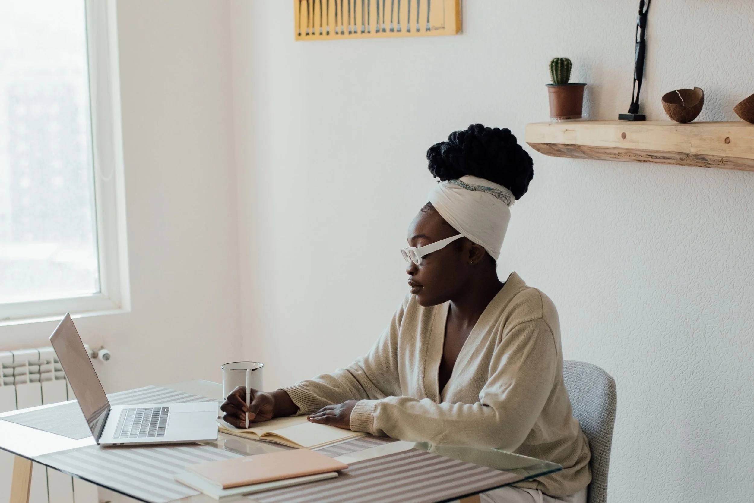 A woman with dark curly hair in a bun, wearing glasses, sitting at a table with a laptop, notebook, cup, and pen, working in a bright room with a window and a wooden shelf with a cactus and decorative objects.
