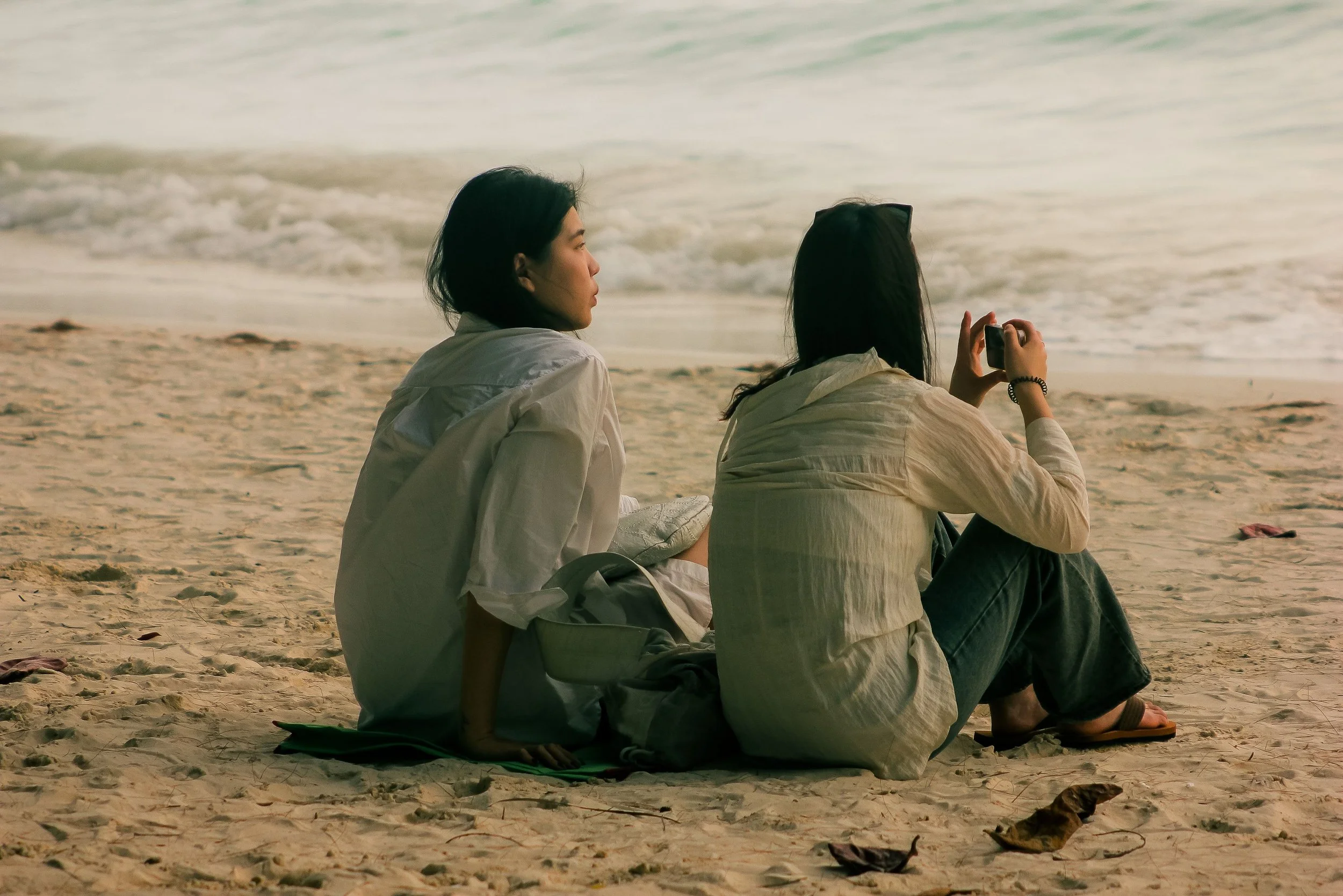 Two young women sitting on a sandy beach near the water, one is using a mobile phone and the other is looking at her.