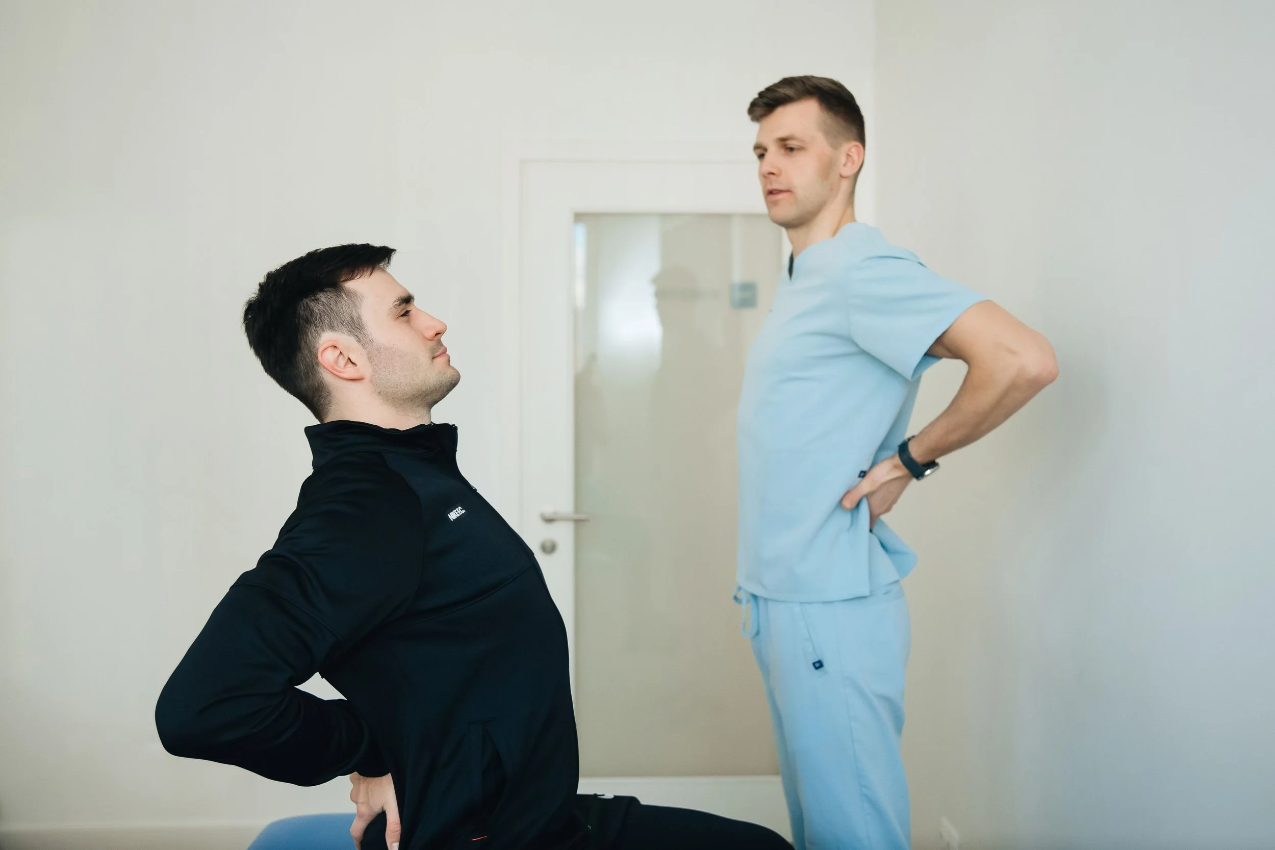 A physical therapist or healthcare professional in light blue scrubs assessing a male patient's posture and back alignment in a clinical setting.