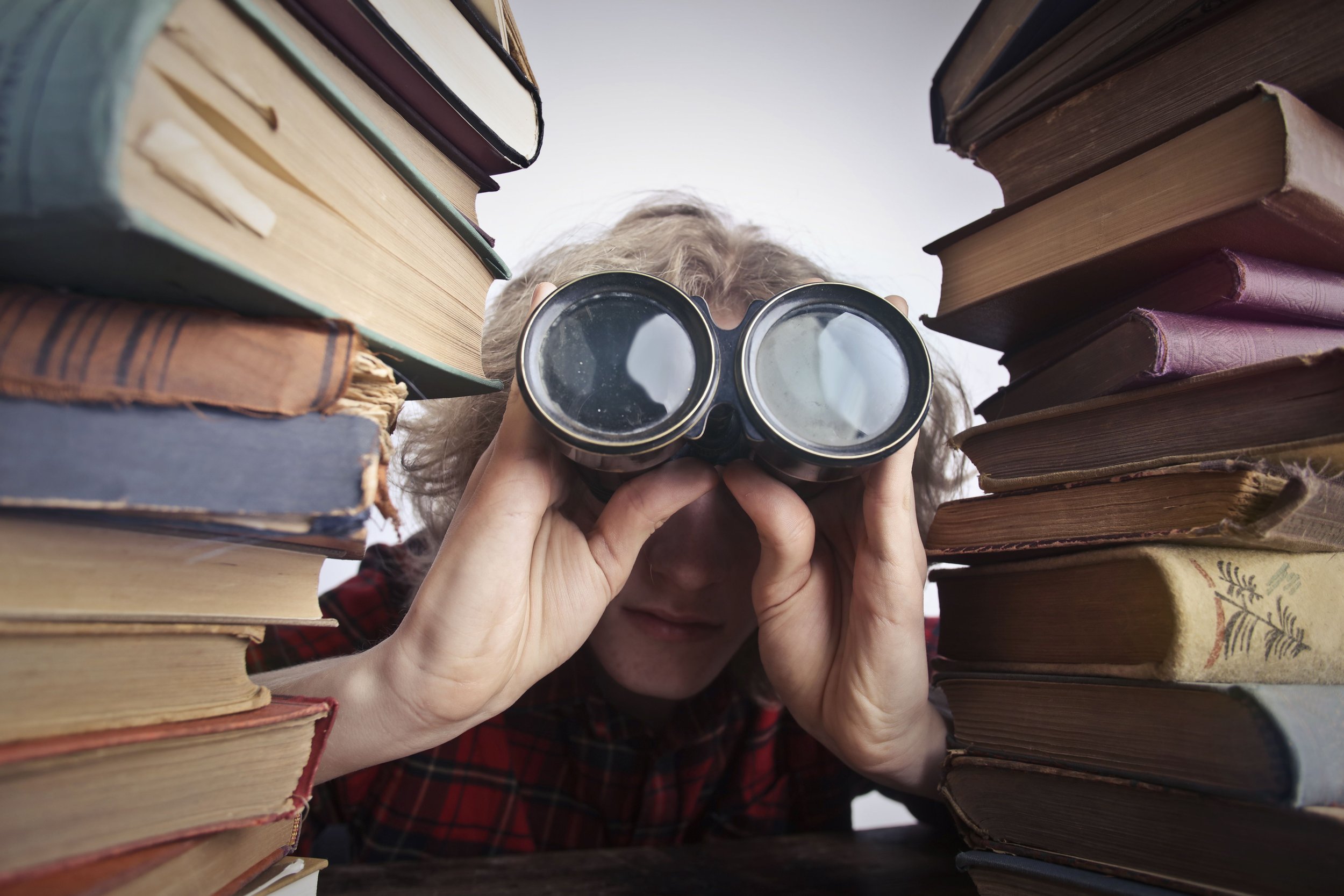 Person with curly hair wearing glasses and a red plaid shirt looking through binoculars between two stacks of old books.