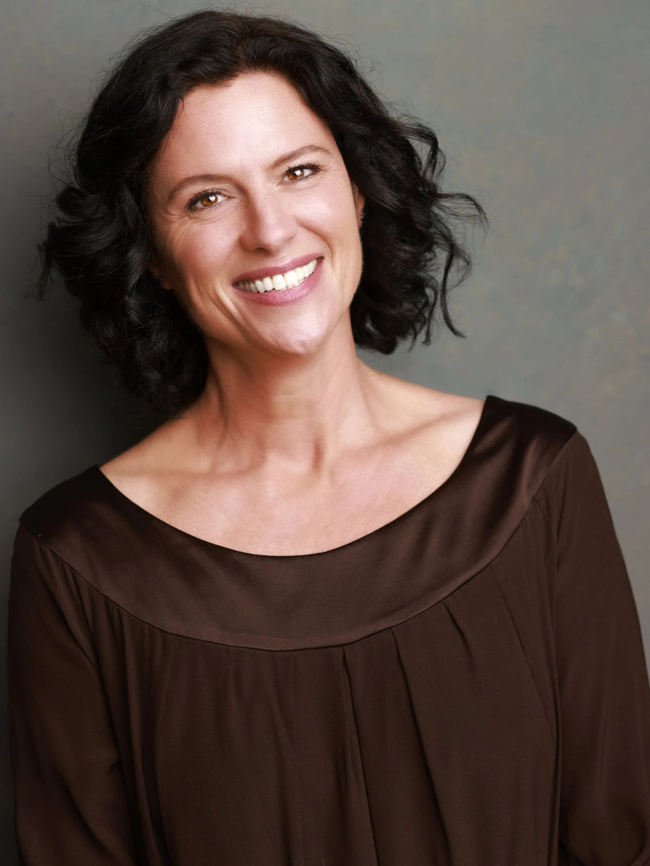 A portrait of a woman with short curly dark hair, smiling, wearing a dark brown dress, against a gray background.