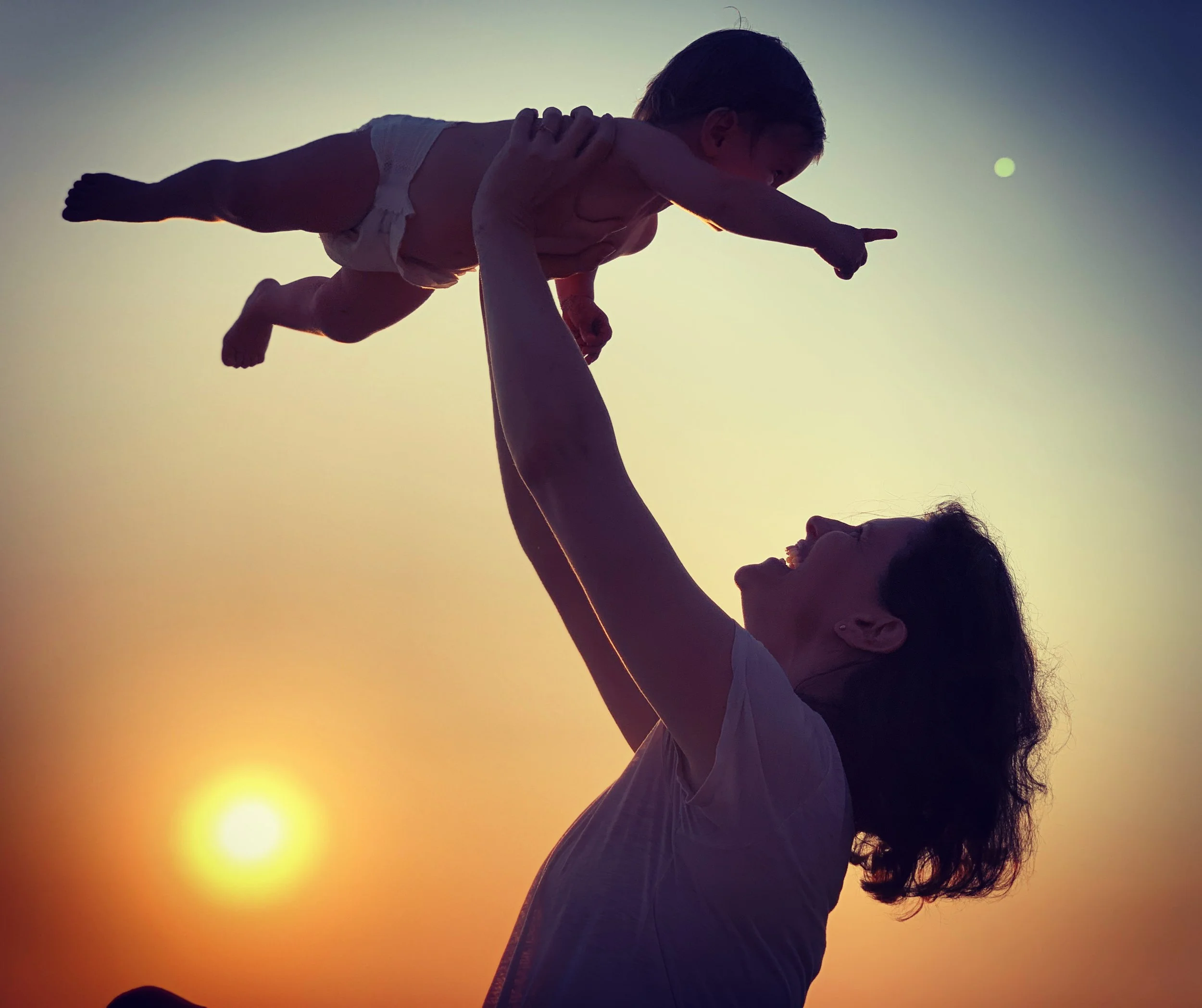 A woman lifts a child in the air during sunset, both facing each other with the sun in the background.