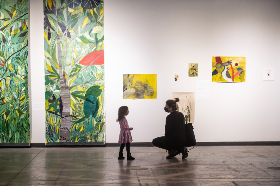 Une adulte masquée observe des œuvres d’art avec une fillette dans une galerie d’exposition.