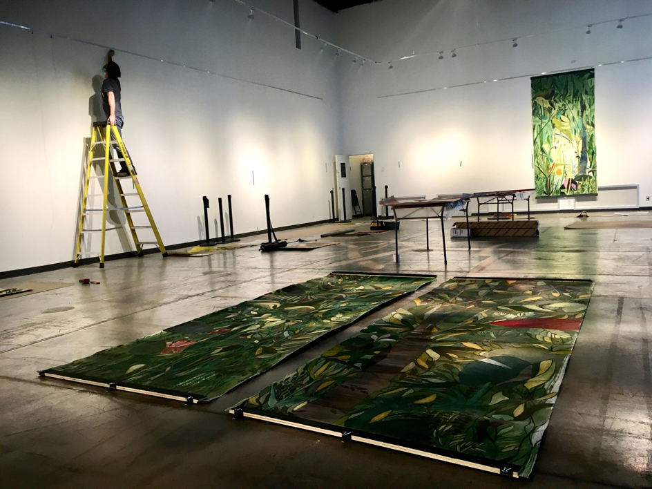 Installation d’une exposition dans une galerie d’art, avec de grandes œuvres végétales au sol et au mur, tandis qu’un technicien travaille sur une échelle.
