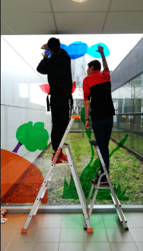 Deux hommes installent des décorations colorées sur une grande baie vitrée, debout sur des escabeaux dans un espace intérieur lumineux.