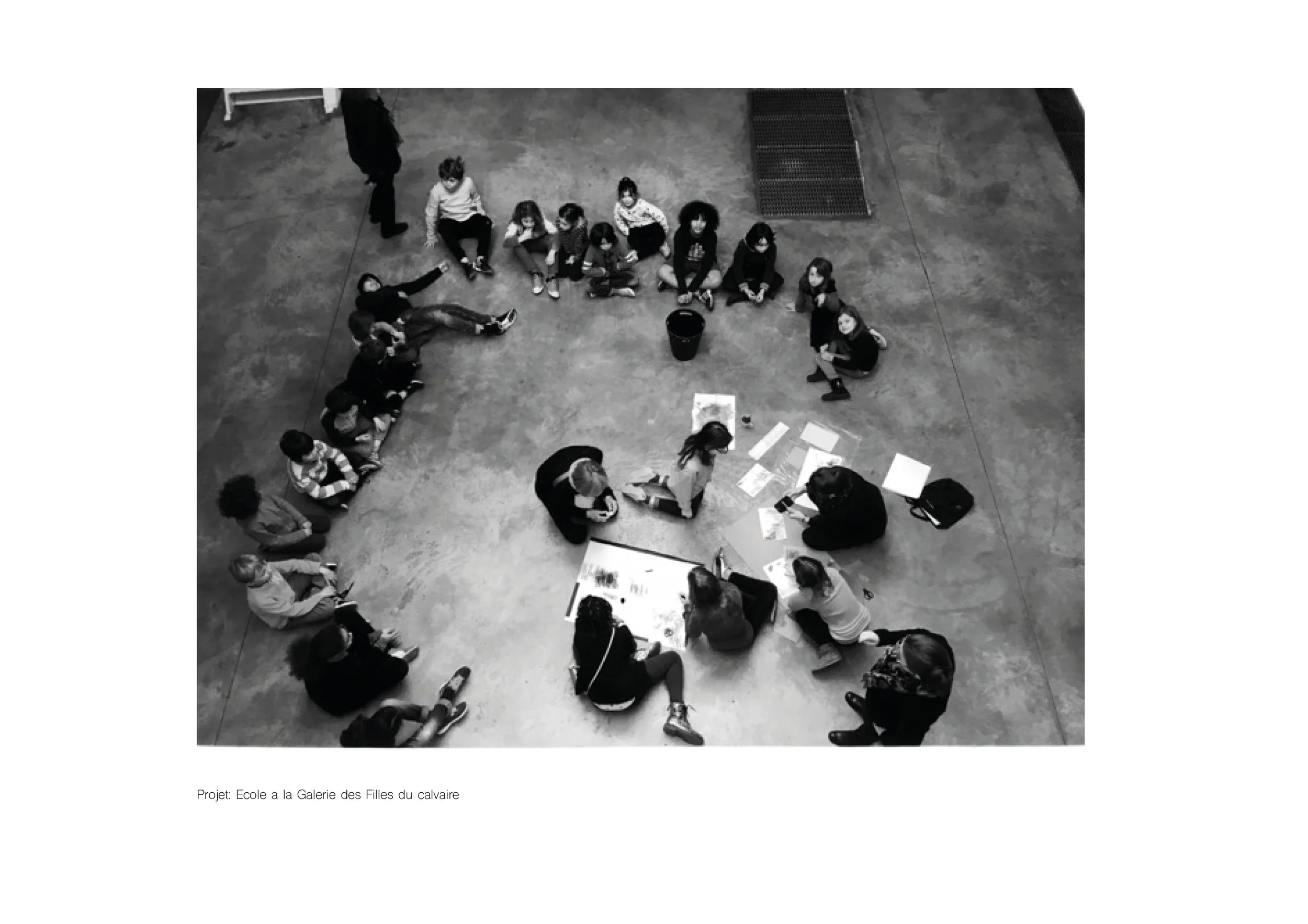 Vue aérienne en noir et blanc d’un groupe d’enfants assis en cercle autour d’activités artistiques au sol dans une galerie.