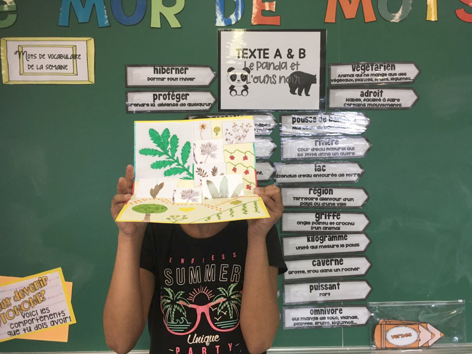 Élève tenant un herbier en papier devant un tableau de classe affichant du vocabulaire français sur la nature et les animaux.
