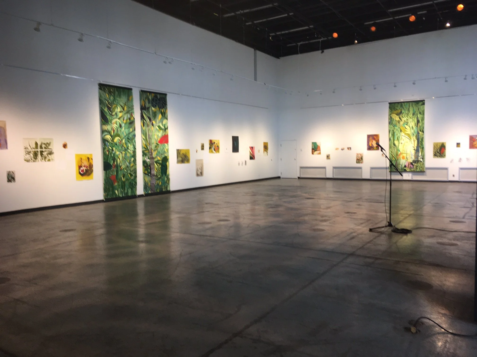 Vue d’une galerie d’art contemporain présentant des peintures colorées, notamment de grandes œuvres végétales, avec un pied de microphone au premier plan.
