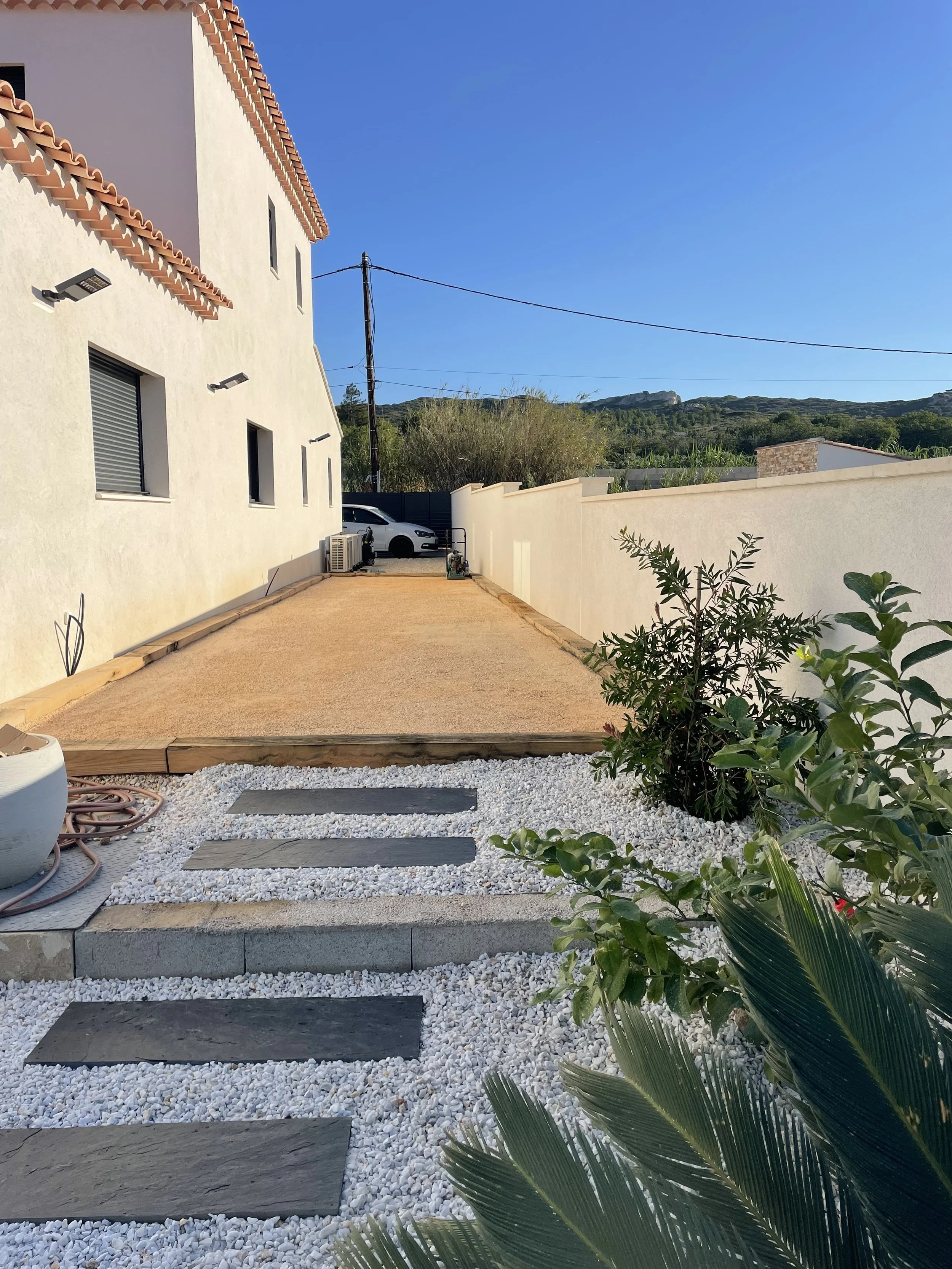 Mediterranean-style home courtyard with white gravel, slate stepping stones, tropical plants, and mountain landscape under a clear blue sky