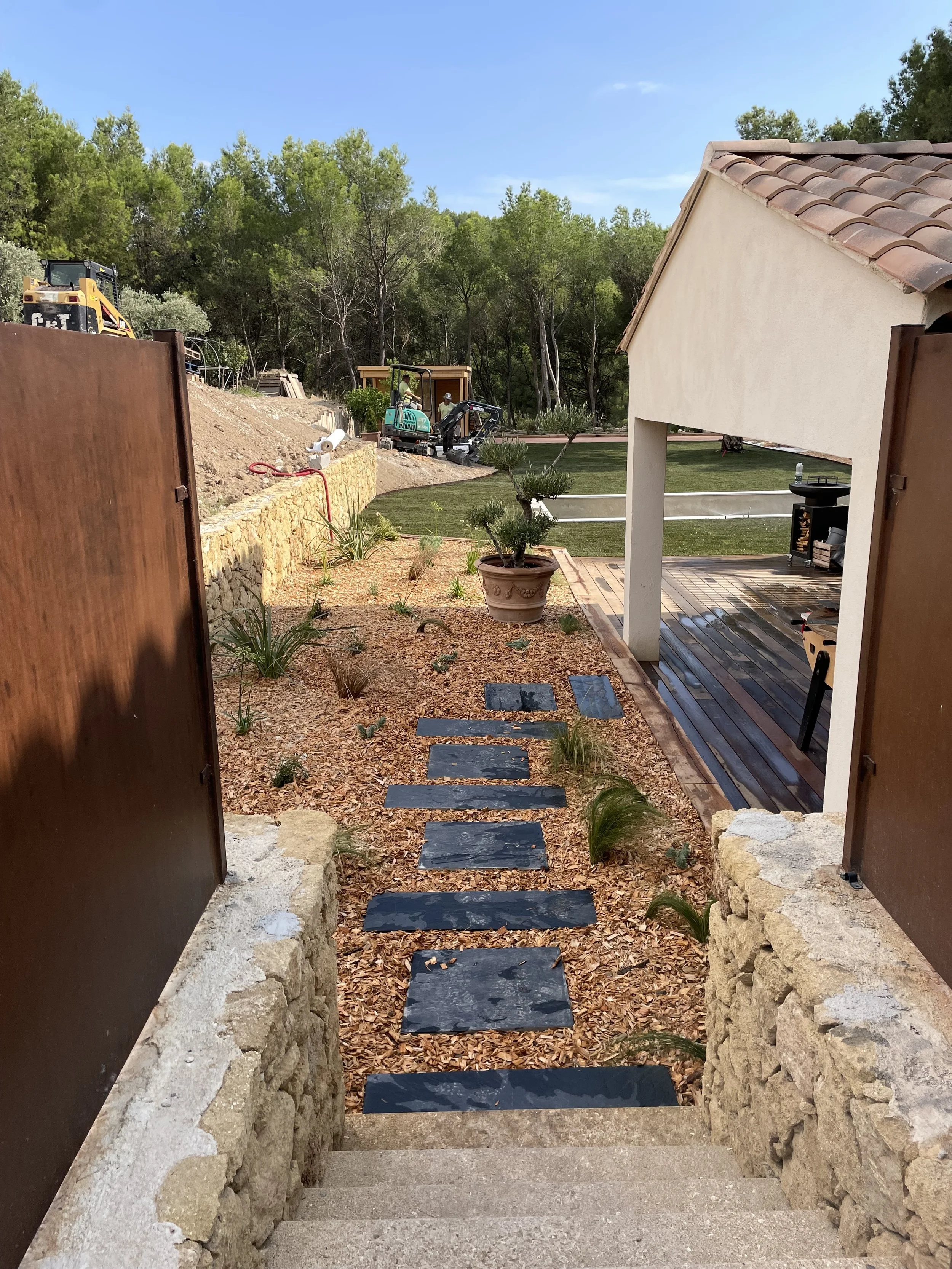 Landscaping construction in a residential backyard with slate stepping stones, stone walls, lawn, tile-roofed house, trees, and excavators.