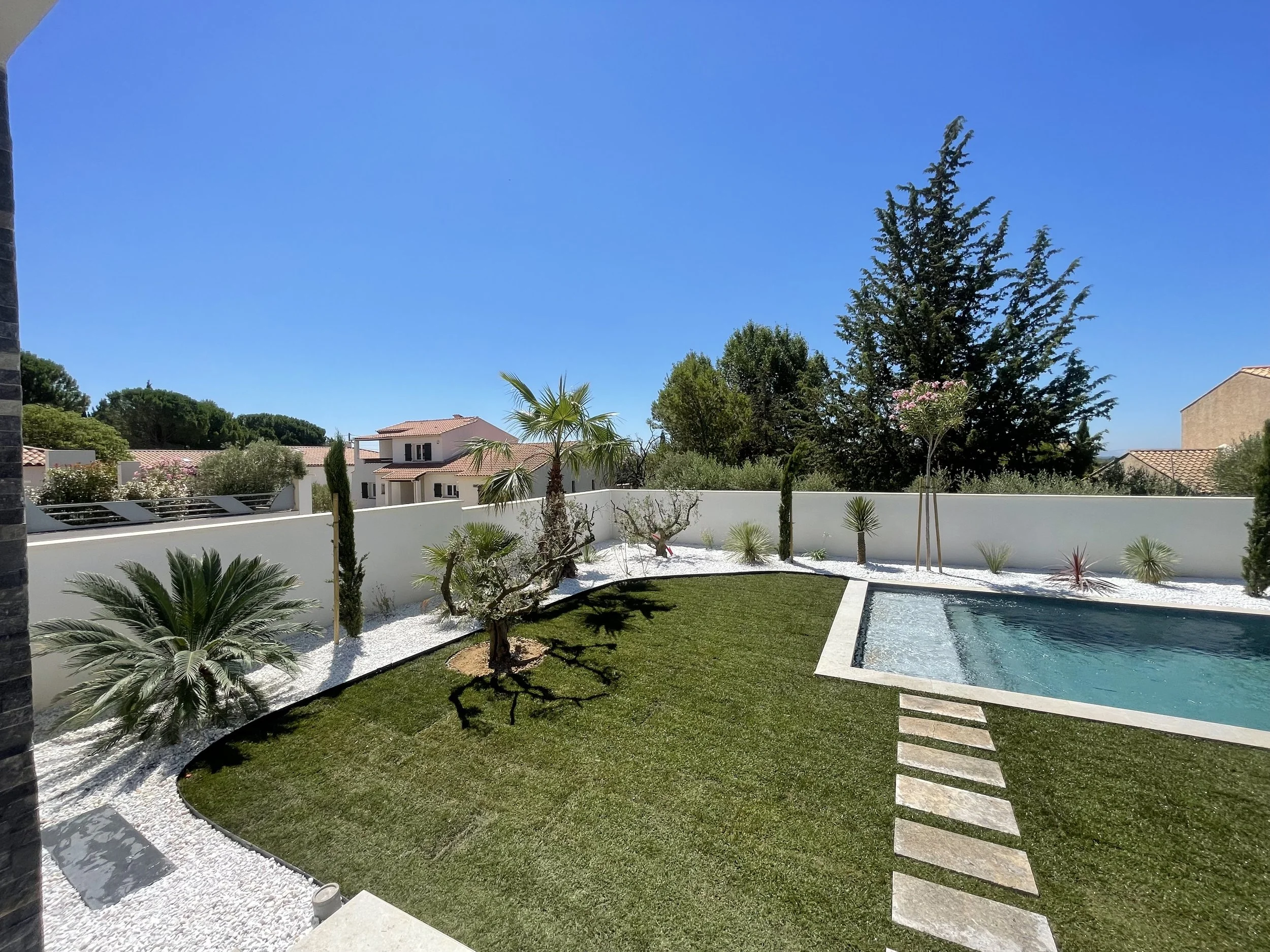 Sunny backyard with a rectangular swimming pool, manicured lawn, palm trees, white gravel landscaping, and Mediterranean-style homes