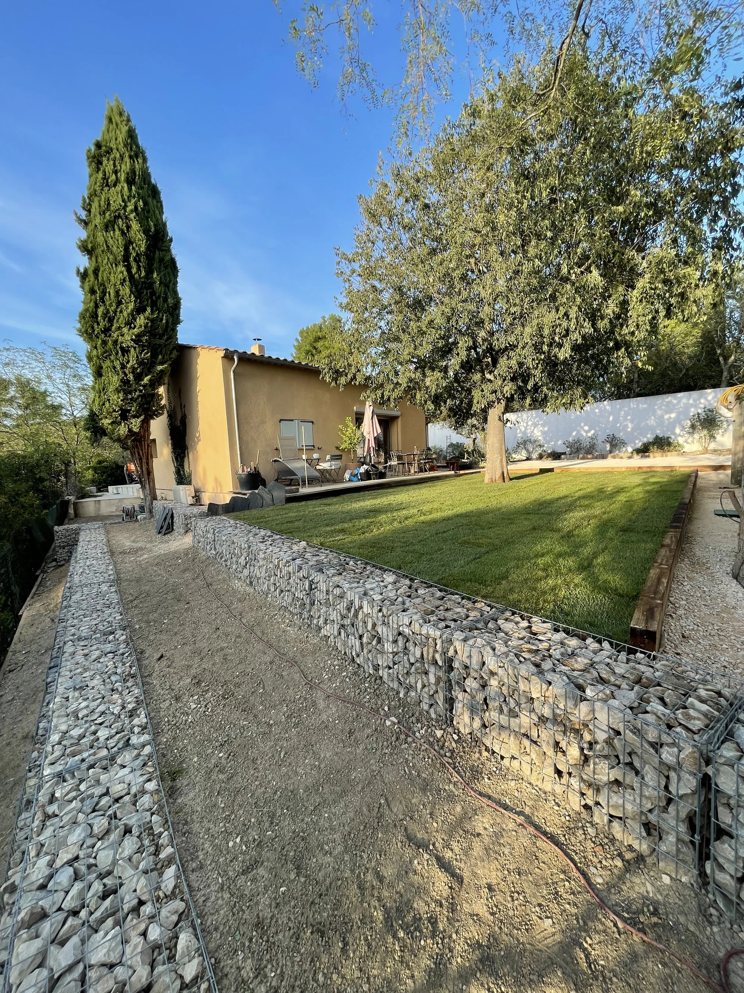 Sunny residential backyard with a manicured lawn, patio furniture, mature trees, a stucco house, and stone-filled gabion landscaping walls.