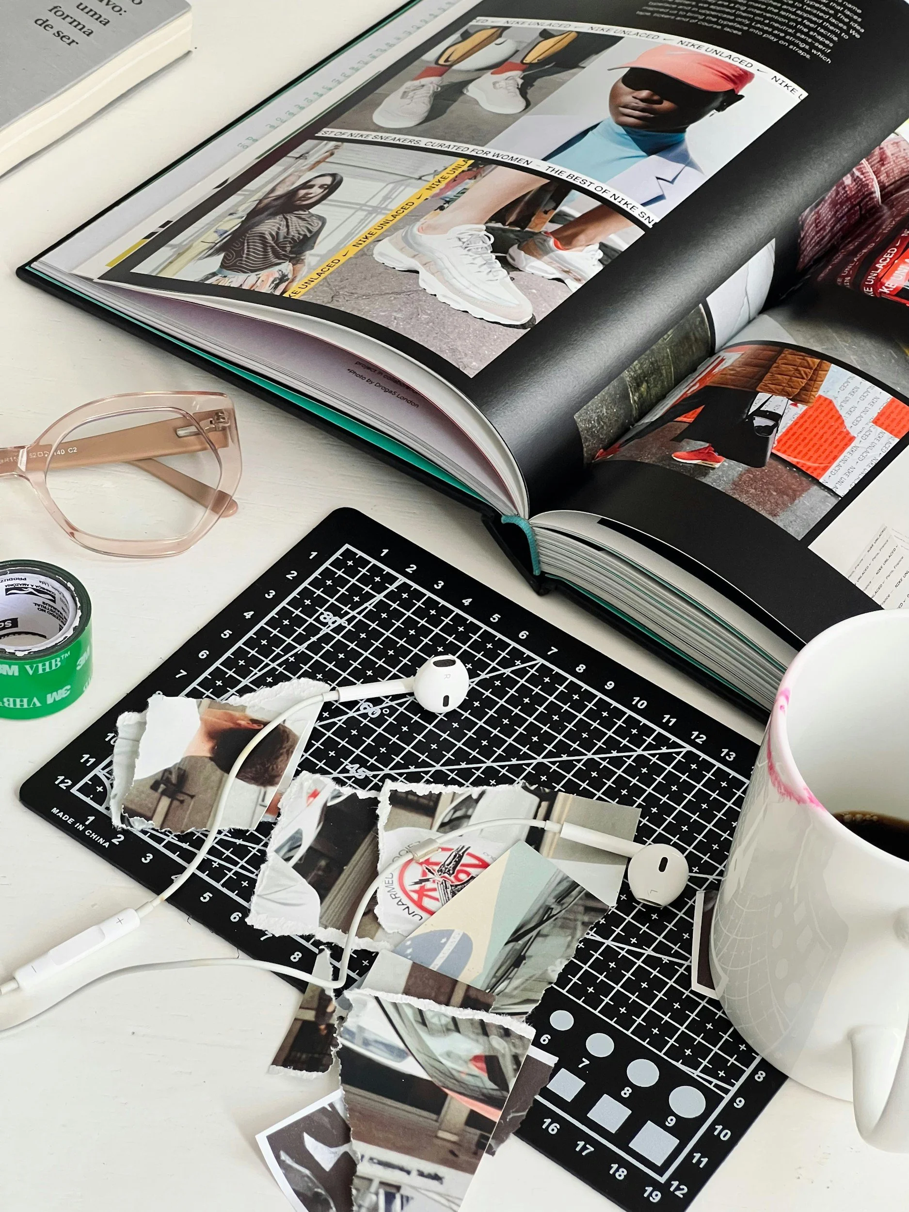 A cluttered desk with torn photographs, a pair of pink glasses, a cutting mat with measurement markings, a roll of tape, earphones, a mug, and an open magazine showing images of people wearing sneakers and casual clothing.
