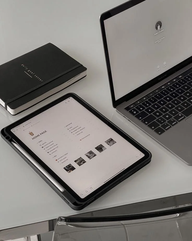 A smartphone on a desk displaying a Pinterest home page, a laptop showing a login screen, and a black notebook labeled 'Business Notebook'.
