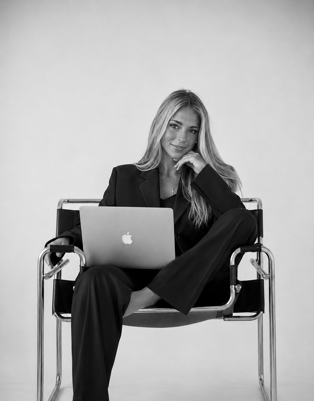 Black and white photo of a woman with long blonde hair sitting on a chair, holding a laptop on her lap, smiling, with her right hand resting on her chin, wearing a dark blazer and pants.