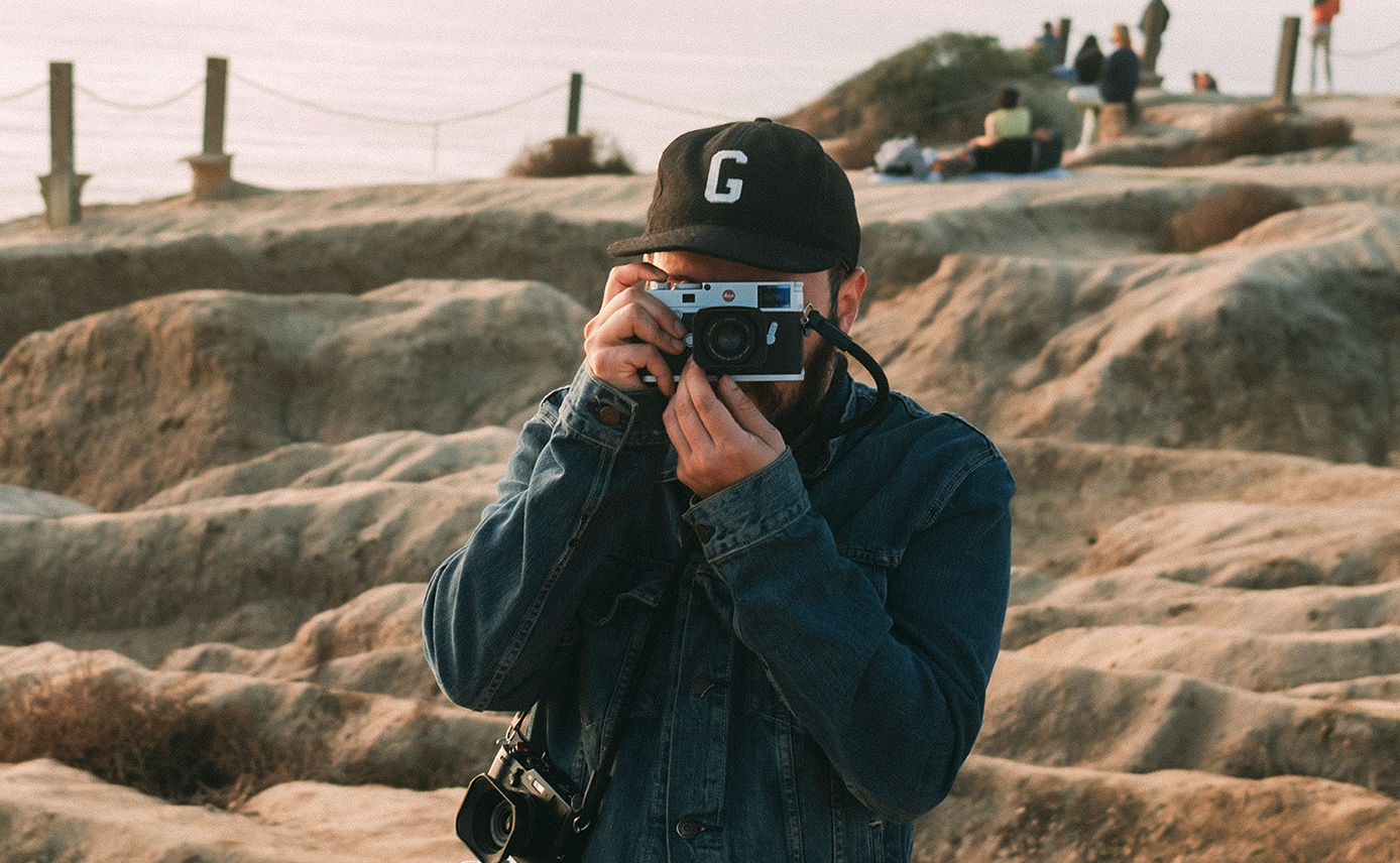 A person wearing a black baseball cap with a white letter 'G' on it, a denim jacket, and holding a camera up to their face, taking a picture on a rocky beach with people sitting and walking in the background during sunset.