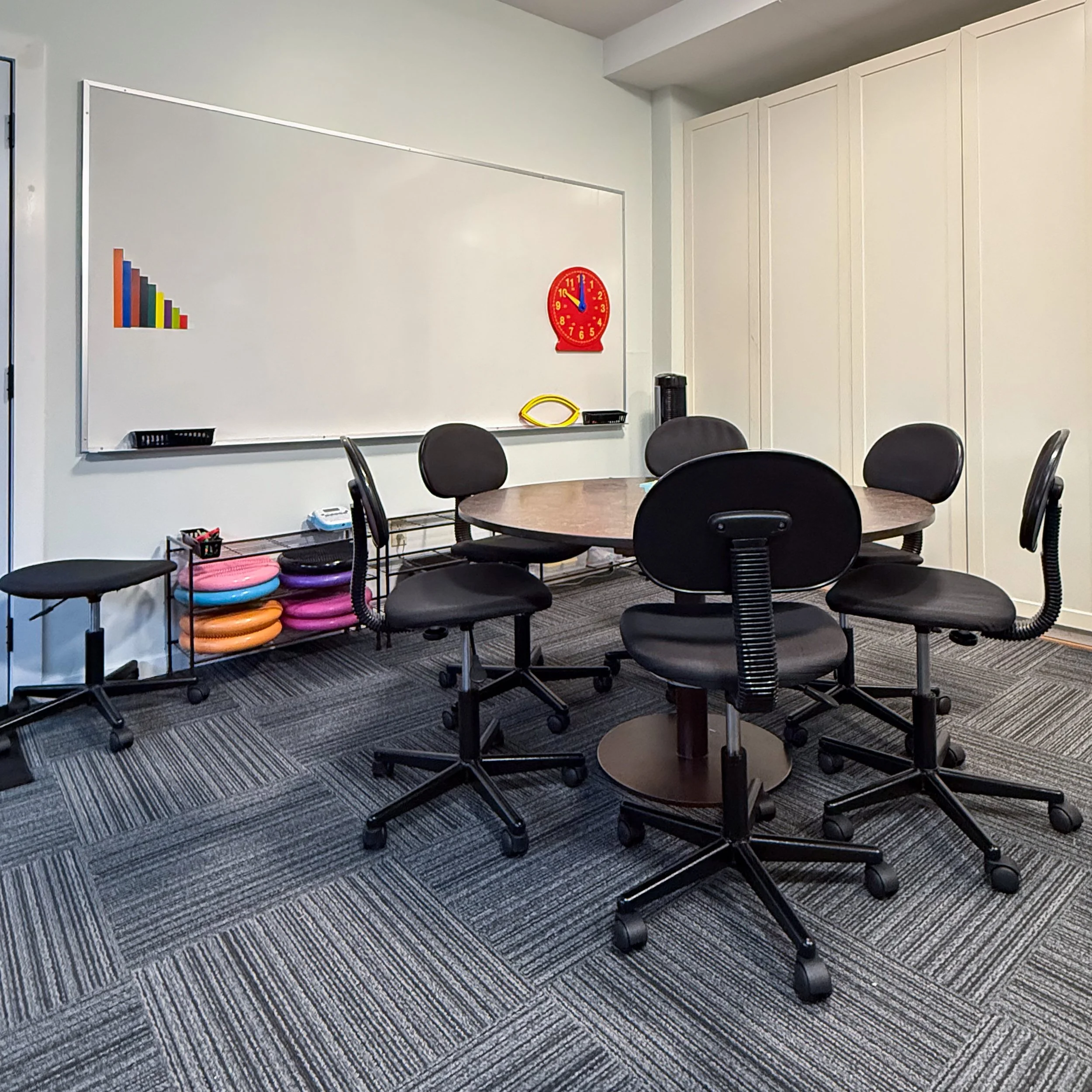 A classroom with a round table and black office chairs around it, a whiteboard, colorful stacking seats, and a wall clock