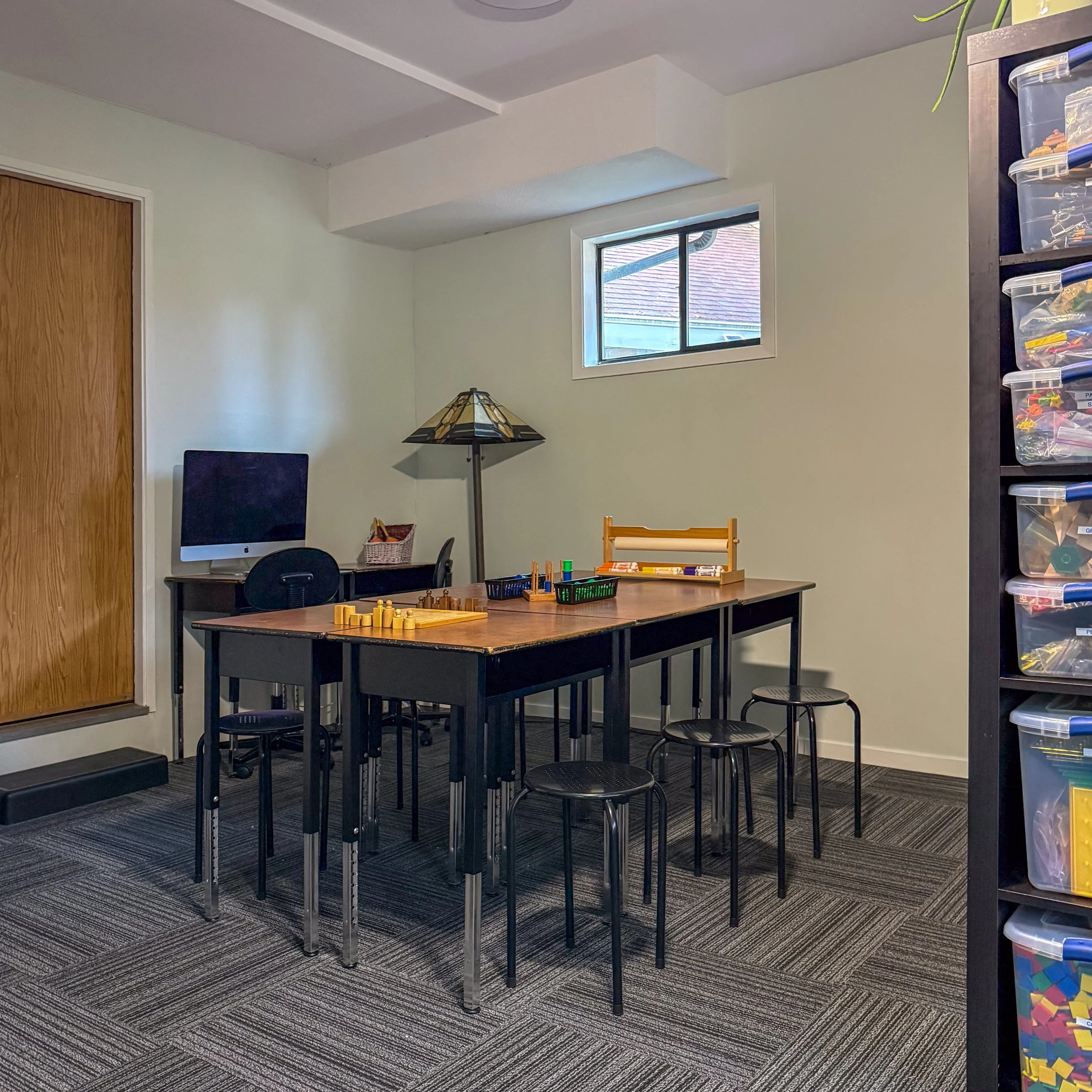 A part of a room with a table, chairs, a computer, a lamp, a basket, and some toys or blocks, with a window and a wooden door in the background, and a storage unit with containers on the right.