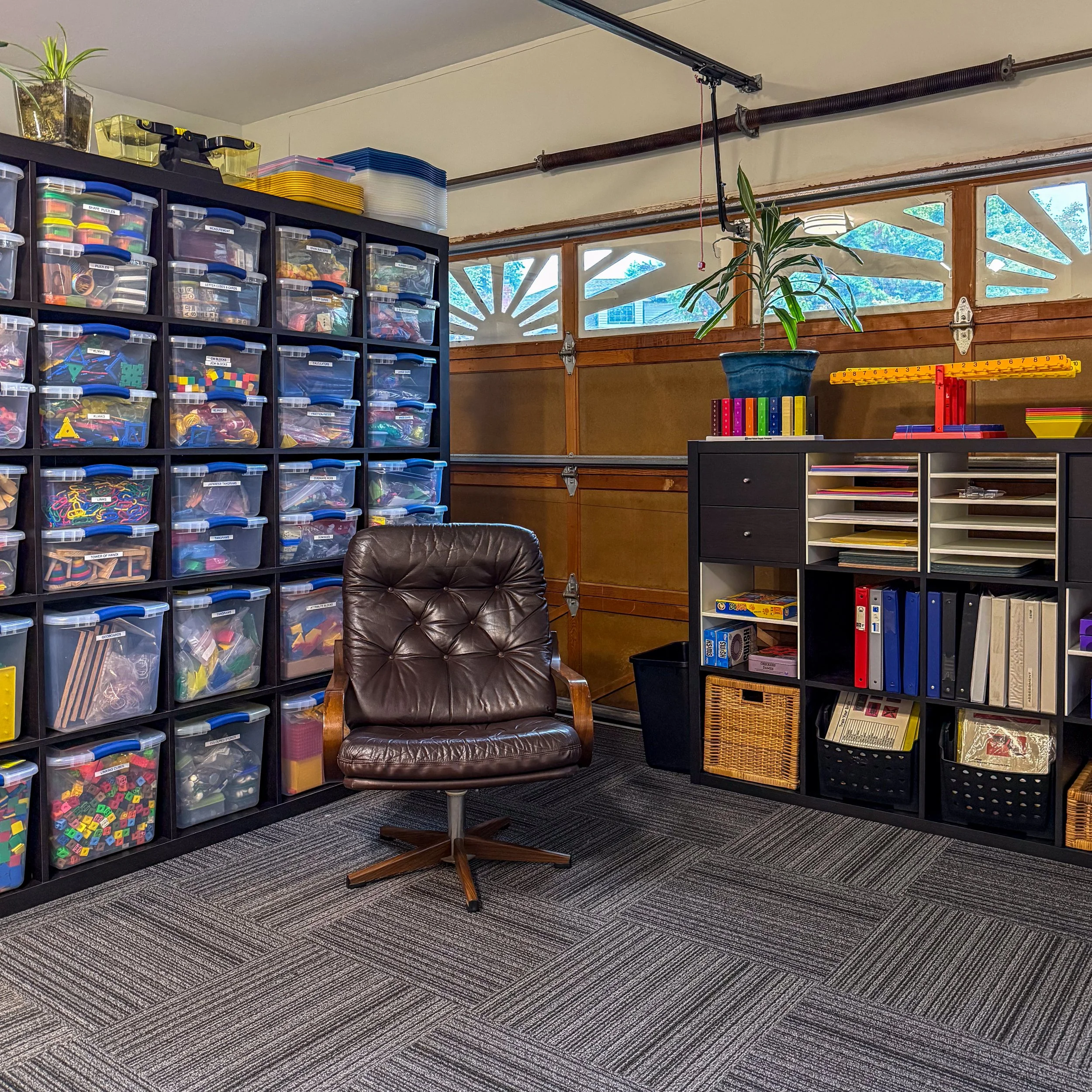A room with storage bins filled with toys or educational materials, a brown leather chair, a small black cabinet, a bookshelf with organized binders, a potted plant, and a garage door with windows showing trees outside.