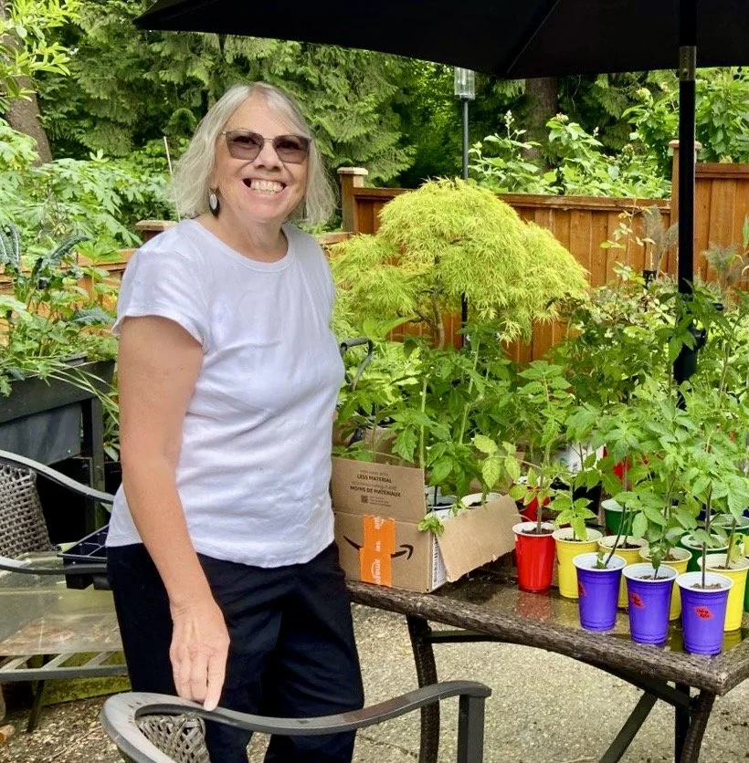 Woman with short gray hair, sunglasses, and a white shirt standing outdoors next to a table with colorful cups and potted plants in a backyard garden.