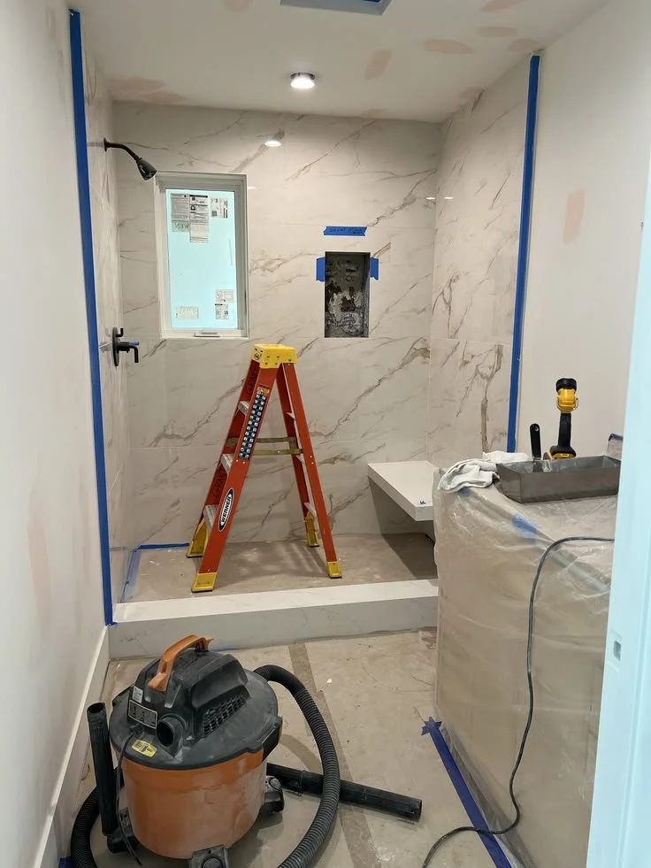 Bathroom under renovation with marble tiled walls, window, step-up shower area, ladder, and construction tools.