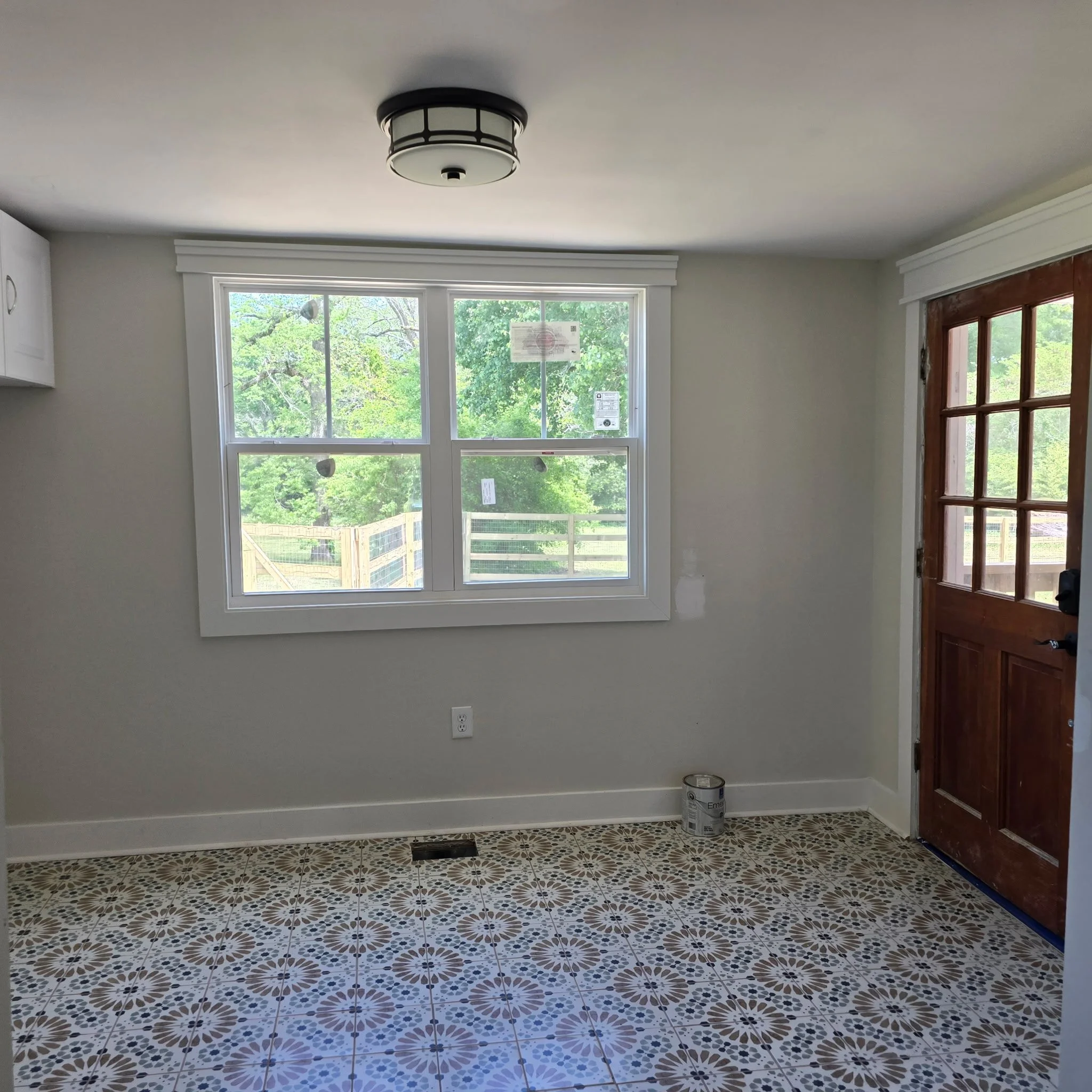 A room under renovation with a large window, patterned tile floor, a white wall, a ceiling light fixture, a closed wooden door, and a paint can on the floor.