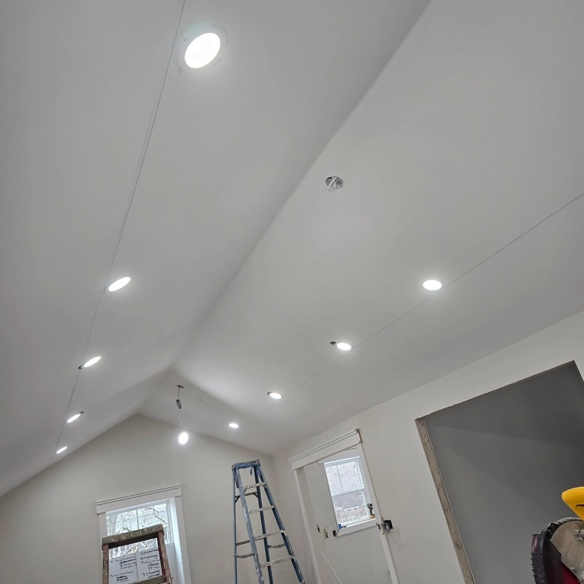 A room under construction or renovation with white walls, multiple ceiling lights, a ladder, and window with blinds. Some tools and materials are visible, indicating ongoing work.