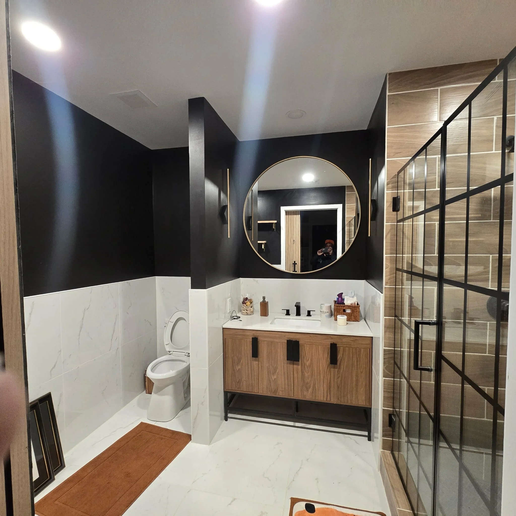 Modern bathroom with black and wood accents, round mirror, toilet, and various toiletries on white marble countertop.