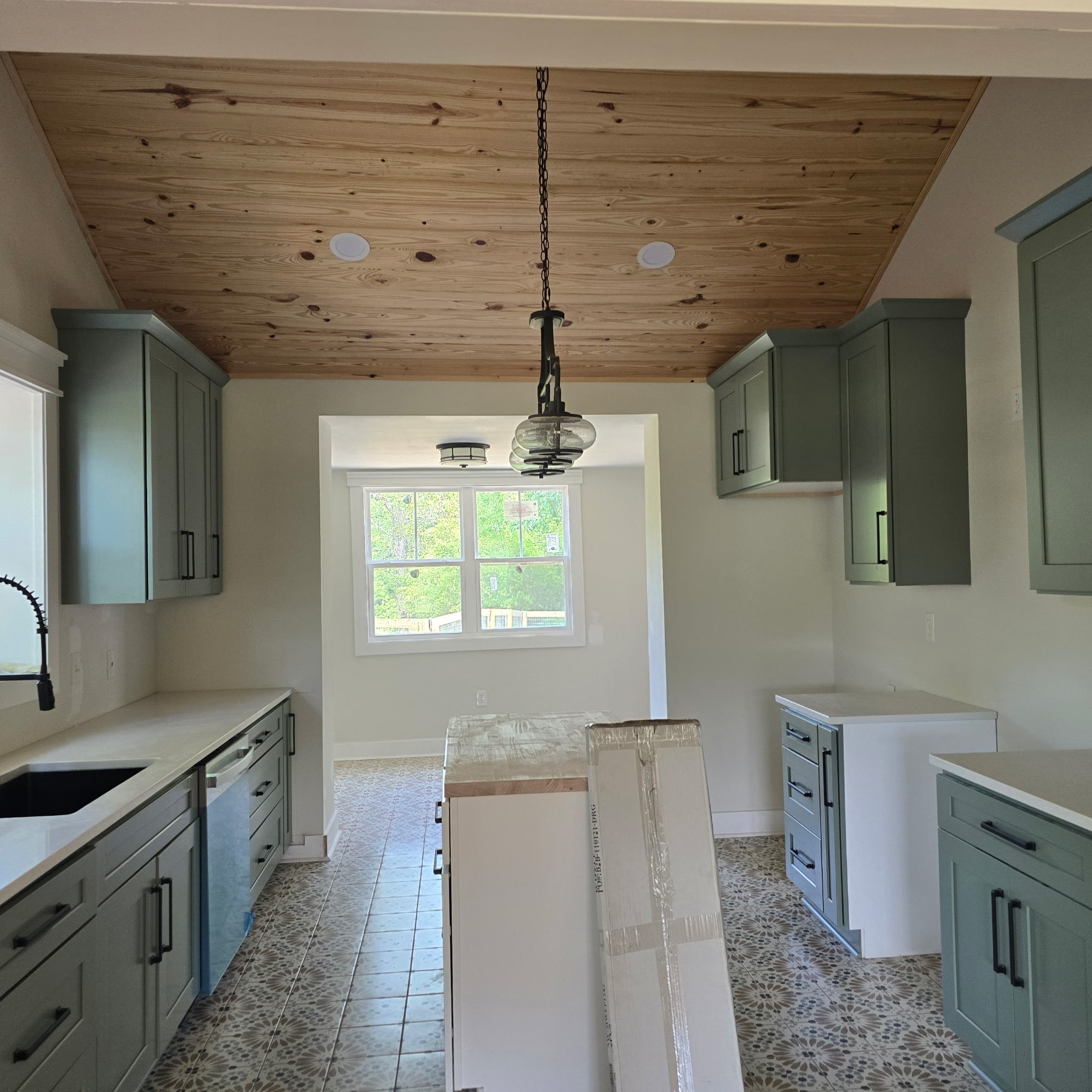 Kitchen with green cabinets, a wooden ceiling, a window showing greenery outside, pendant lighting, and packaging boxes on the floor.