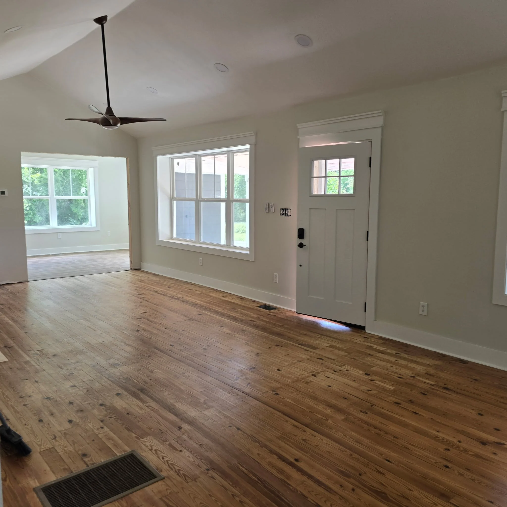 A room with wooden floors, large windows, a ceiling fan, and a white door with a knob and lock. The room appears to be under renovation or construction.