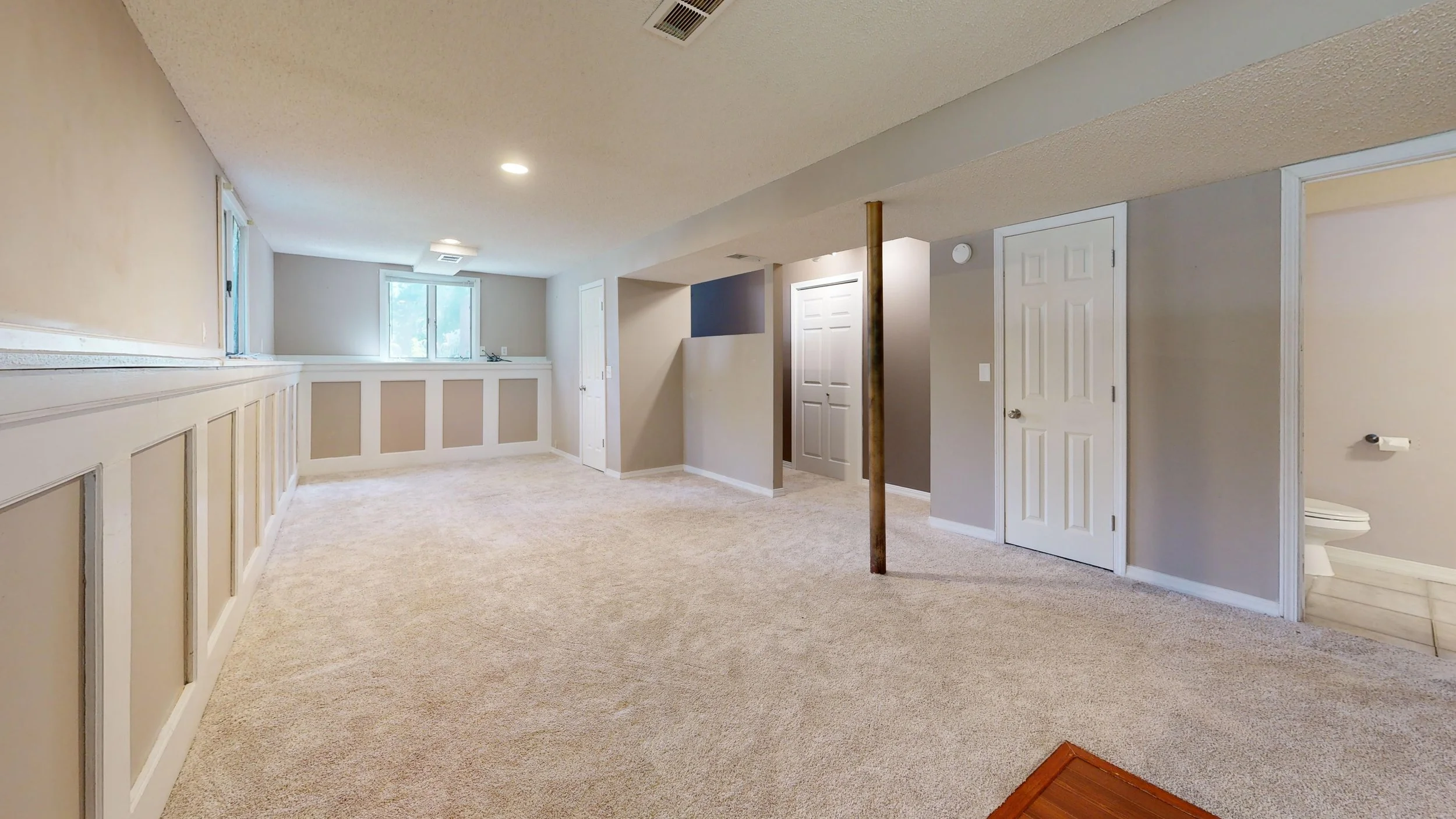 Interior of a basement with beige carpeting, white and gray walls, multiple white doors, a small bathroom with toilet partially visible, windows letting in natural light, and a ceiling vent.