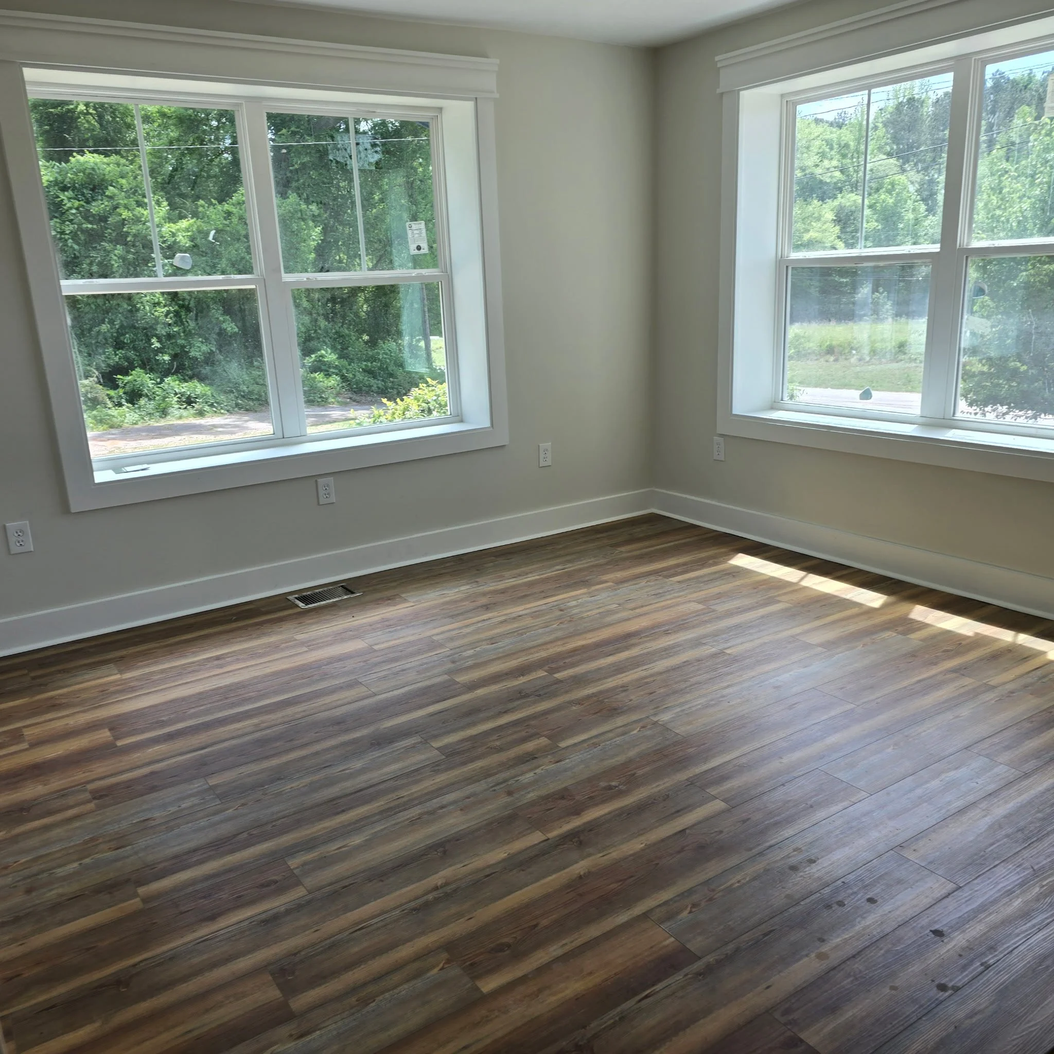 A room with three large windows showing green trees outside, white walls, and dark hardwood flooring.