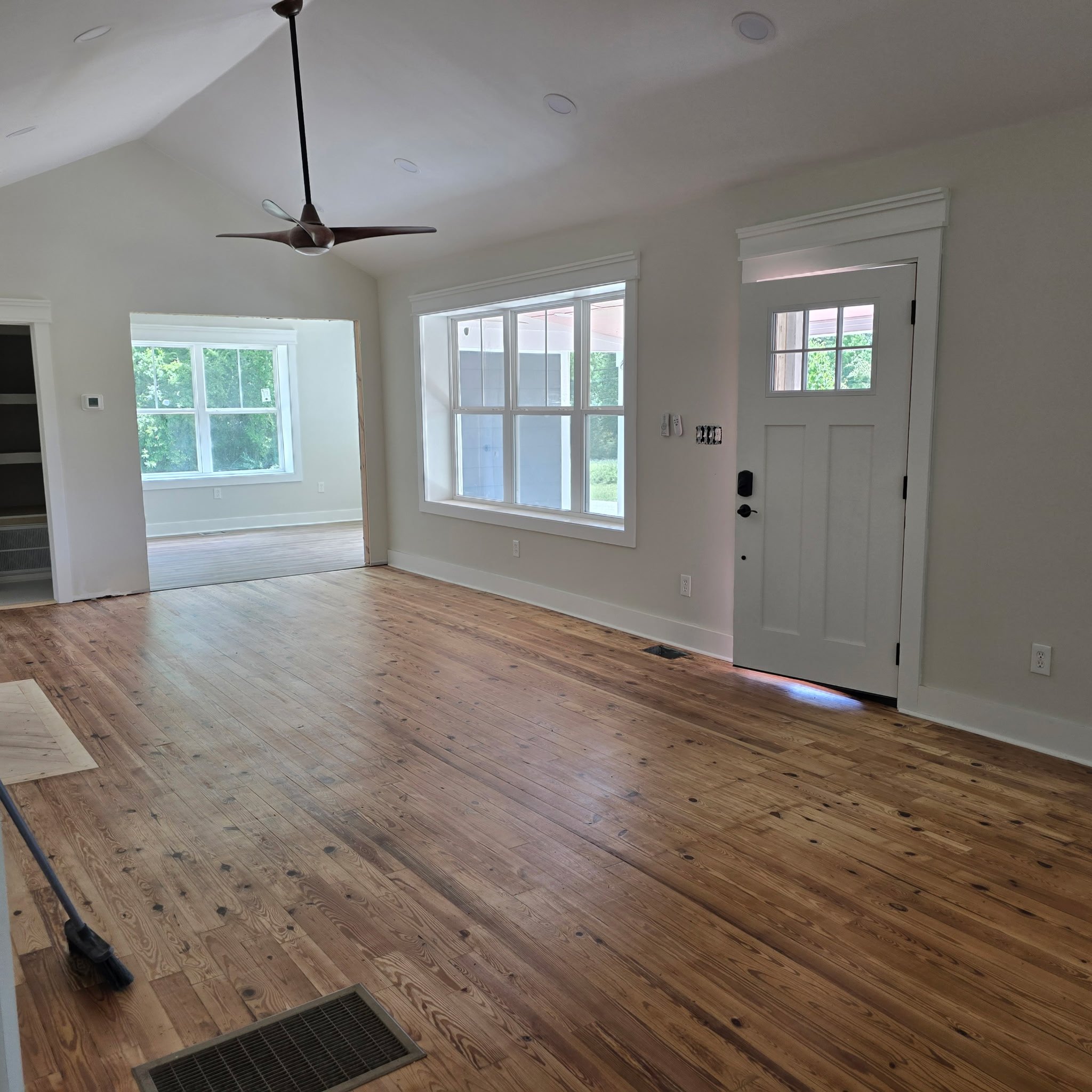 Interior of a house with light-colored walls, large windows, and hardwood floors, with a ceiling fan and an air vent visible.