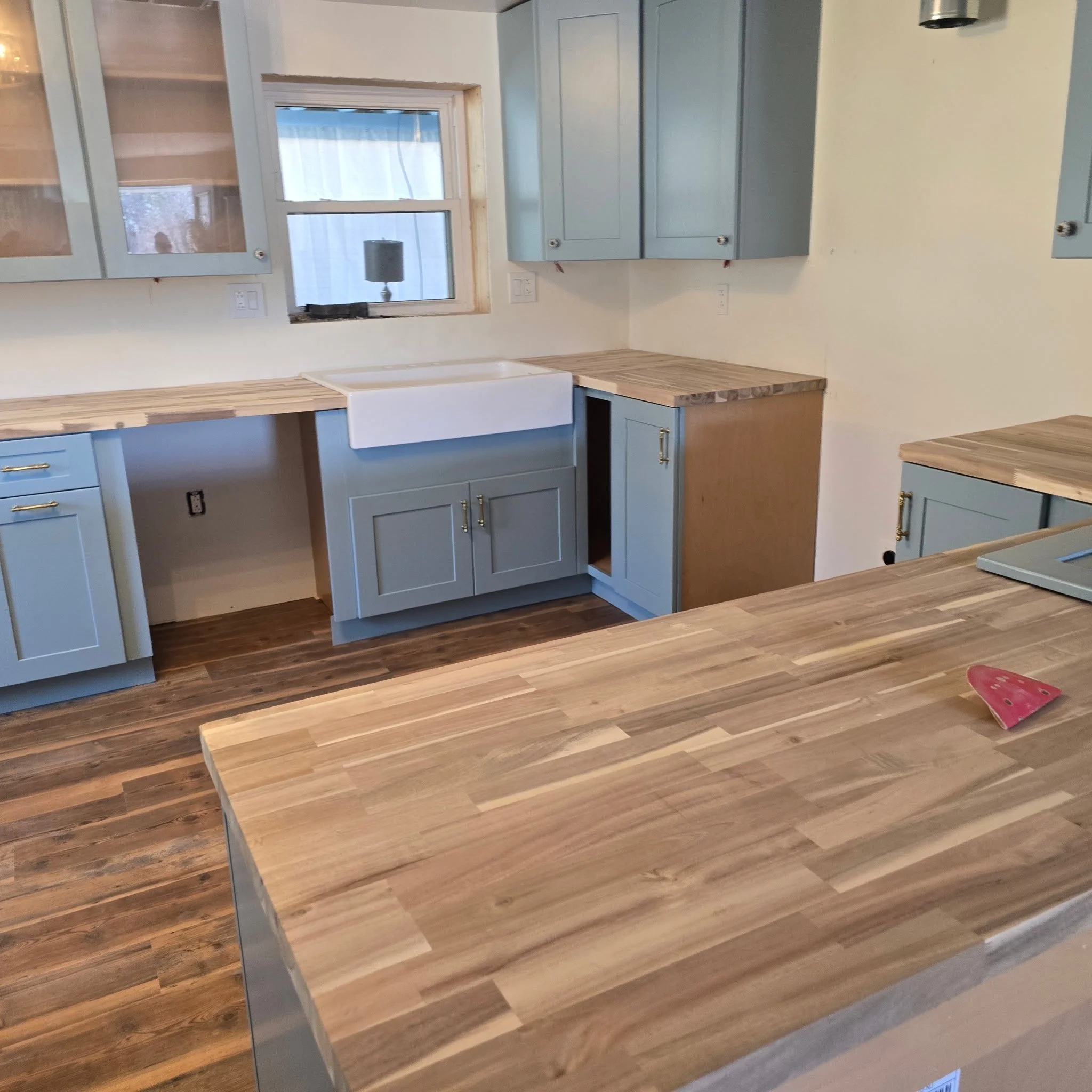 A kitchen with light blue cabinets, wooden countertops, a white farmhouse sink, a window above the sink, light wood flooring, and a few small objects including a pink skateboard on the floor.