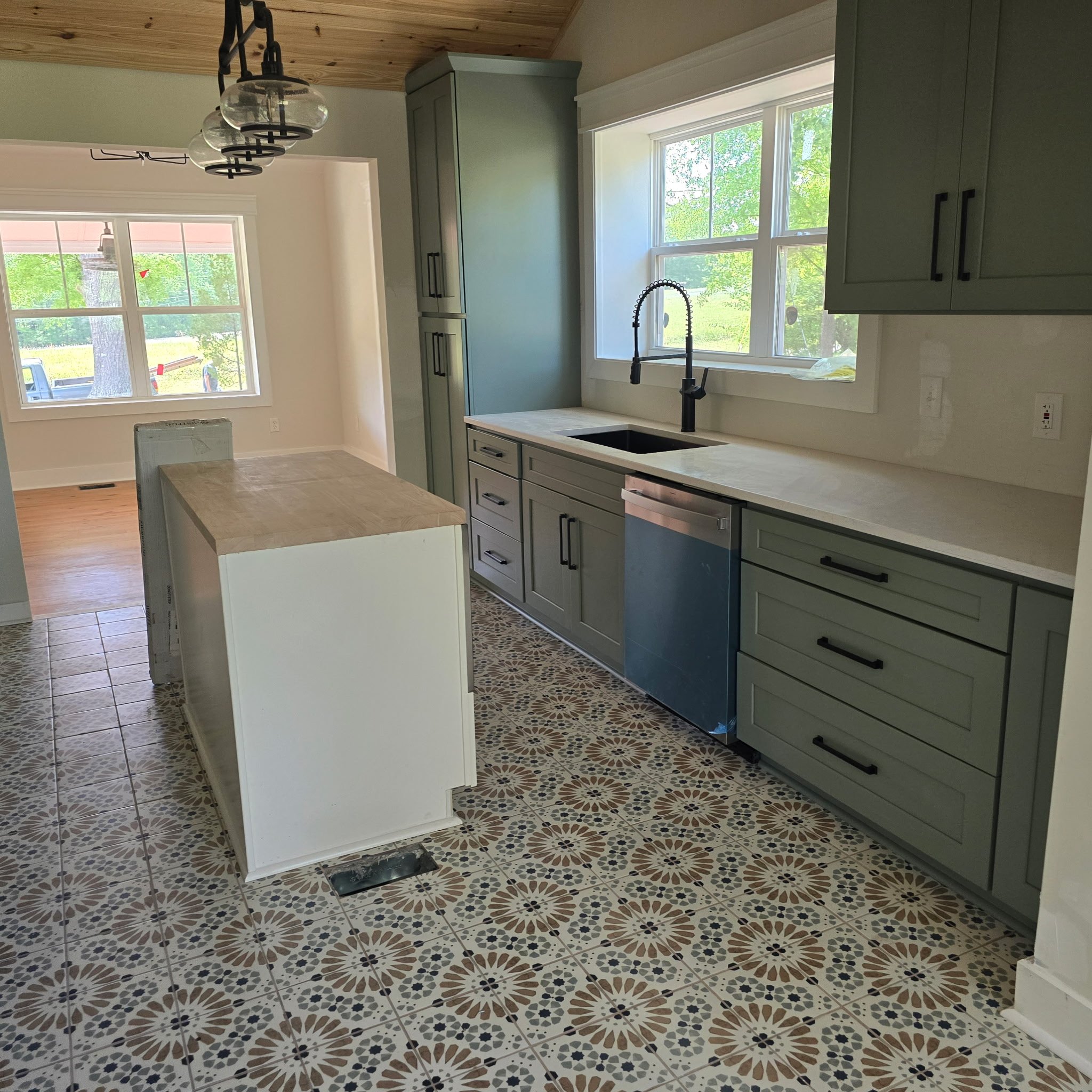 Kitchen with green cabinets, white countertops, large windows showing green trees outside, patterned tile floor, and a hanging light fixture.