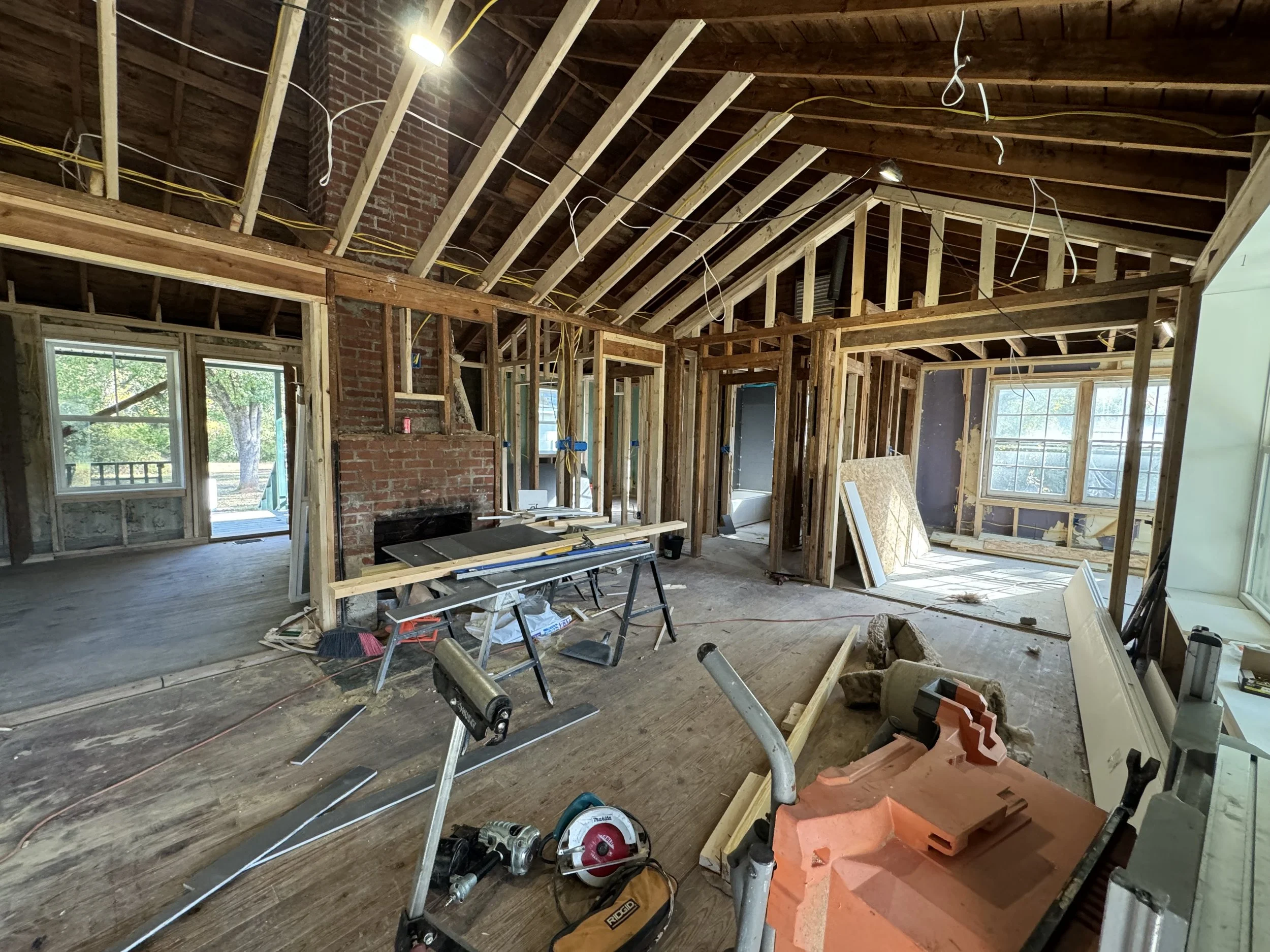The interior of a house under construction with exposed wooden framing, a brick chimney, construction tools, and scattered materials.