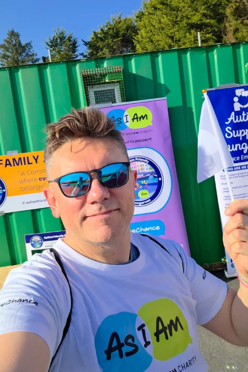 Man wearing sunglasses and a white T-shirt taking a selfie at an outdoor autism awareness event, with colorful posters and flags behind him.