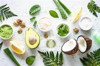 Arrangement of natural skincare ingredients including aloe vera, coconut, lemon, walnuts, green herbs, and various powders on a white background.