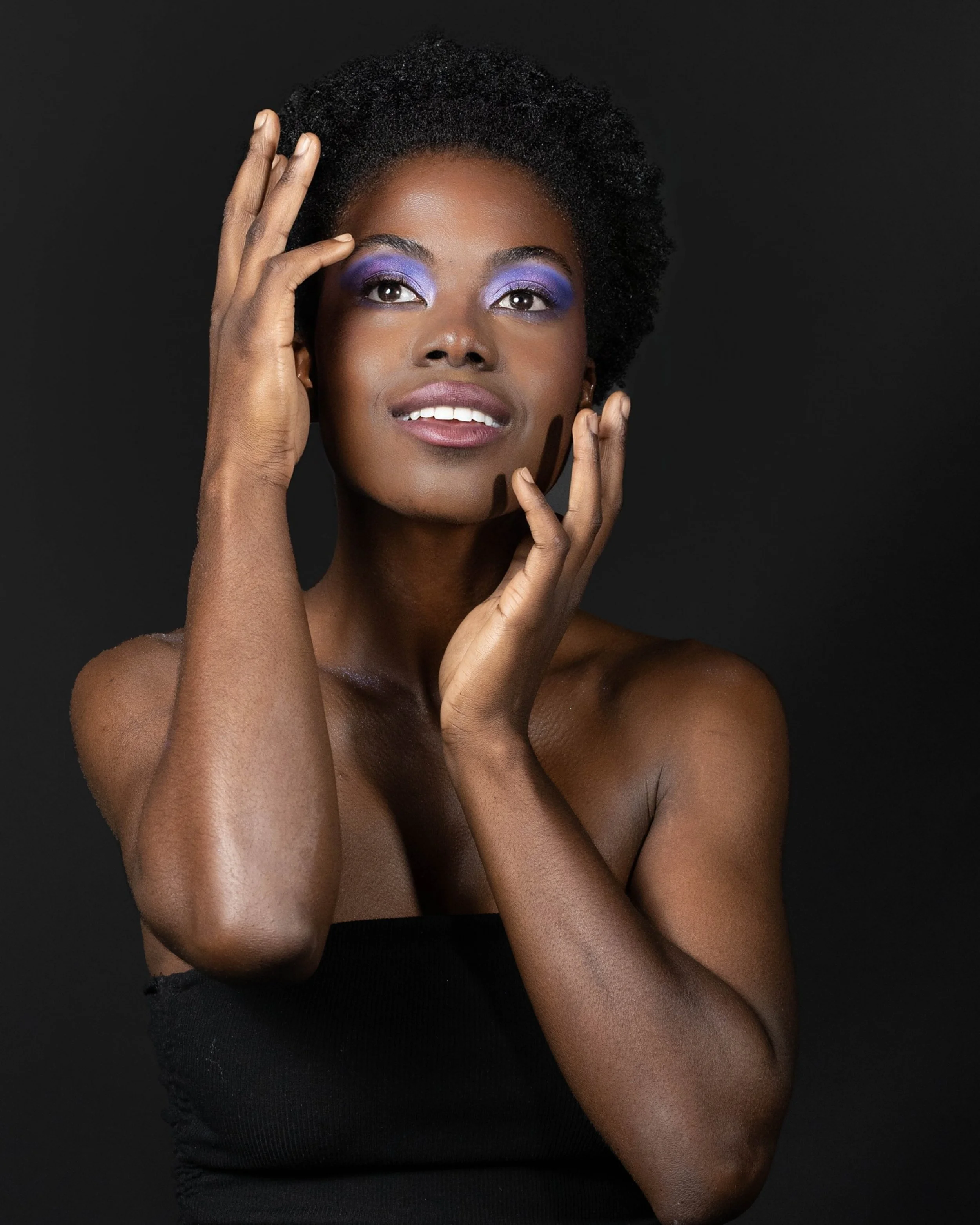 A portrait of a woman with dark skin and short curly black hair, wearing purple eyeshadow and lipstick, with her hands gently touching her face against a black background.
