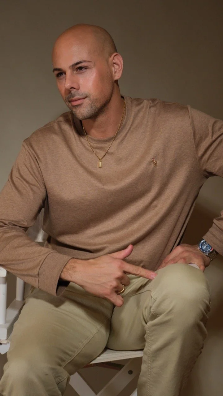A man with a shaved head and light beard, wearing a beige long-sleeve shirt, beige pants, a gold necklace, a wristwatch, and a ring, sitting on a chair indoors with a plain wall background, pointing downward with his right hand.