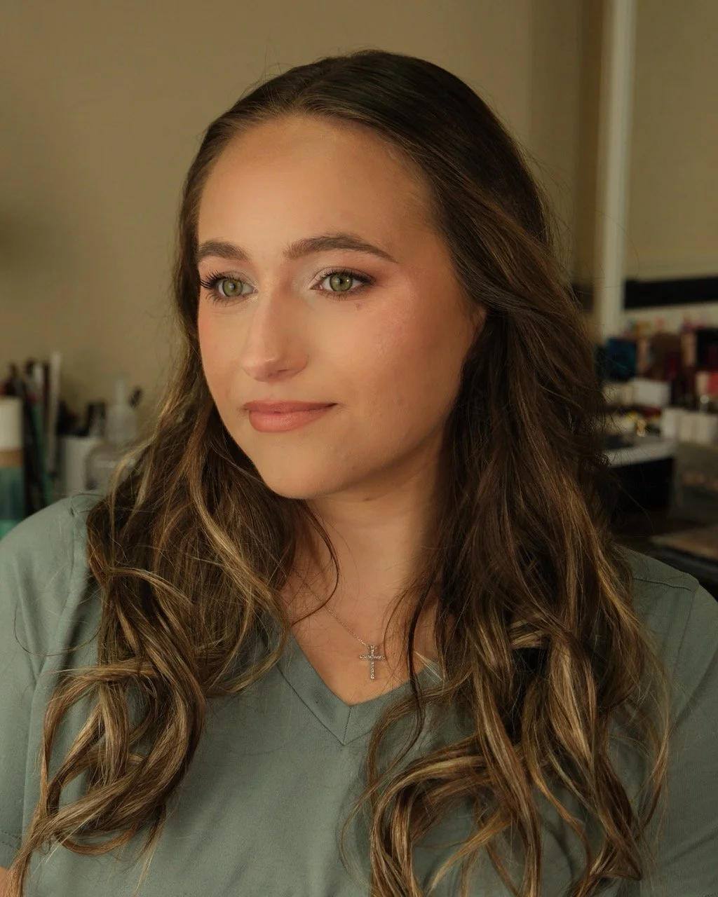 A young woman with long, wavy brown hair and green eyes, wearing a gray V-neck shirt and a silver cross necklace, looking slightly to her left in a softly lit room with a blurred background.